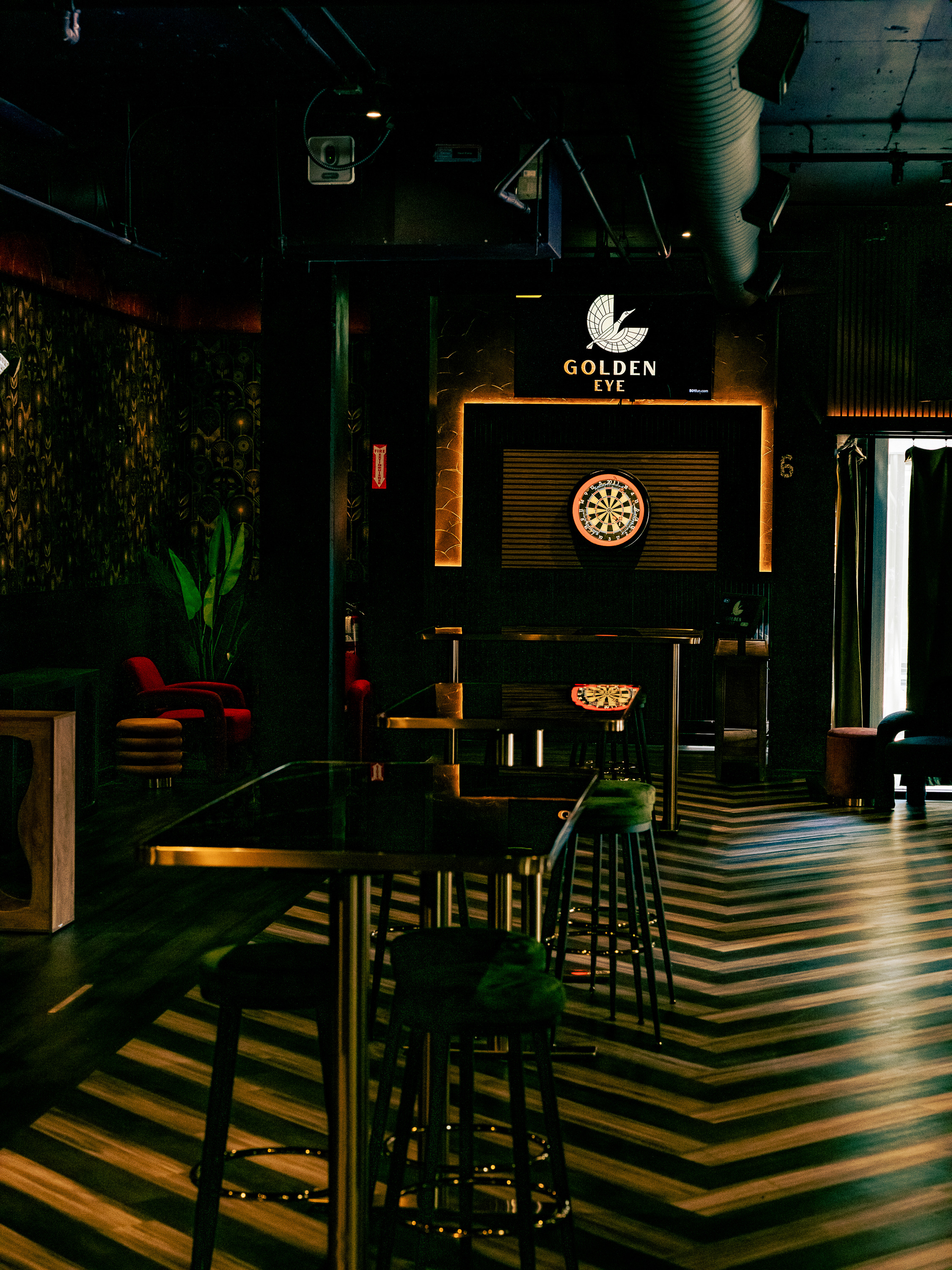 The image shows a dimly lit bar with high tables and stools. A dartboard is mounted on the back wall, and the floor has a zigzag pattern.