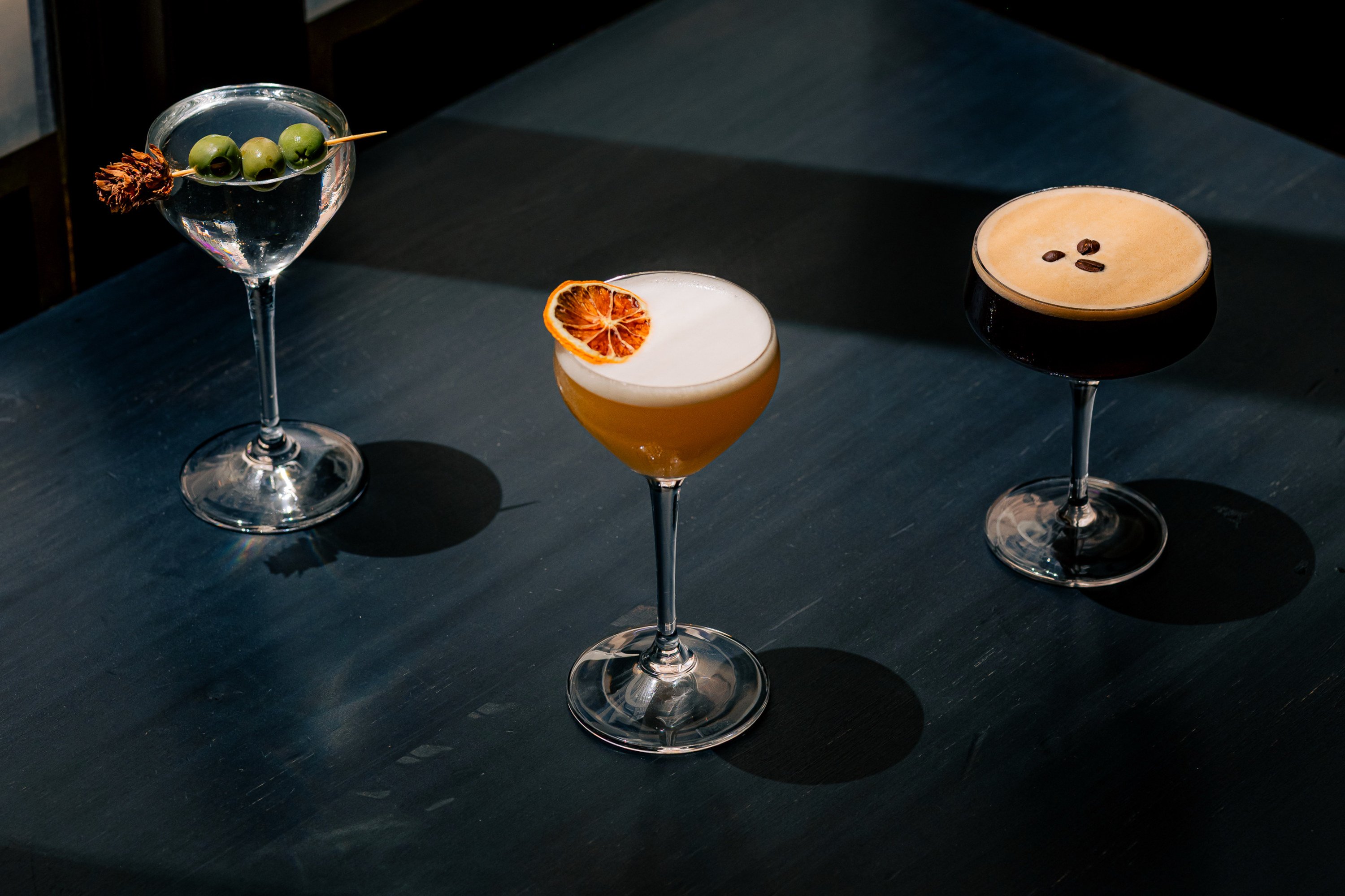 Three elegant cocktails are on a dark table: a martini with olives, a citrus cocktail with foam and slice, and a dark drink garnished with coffee beans.