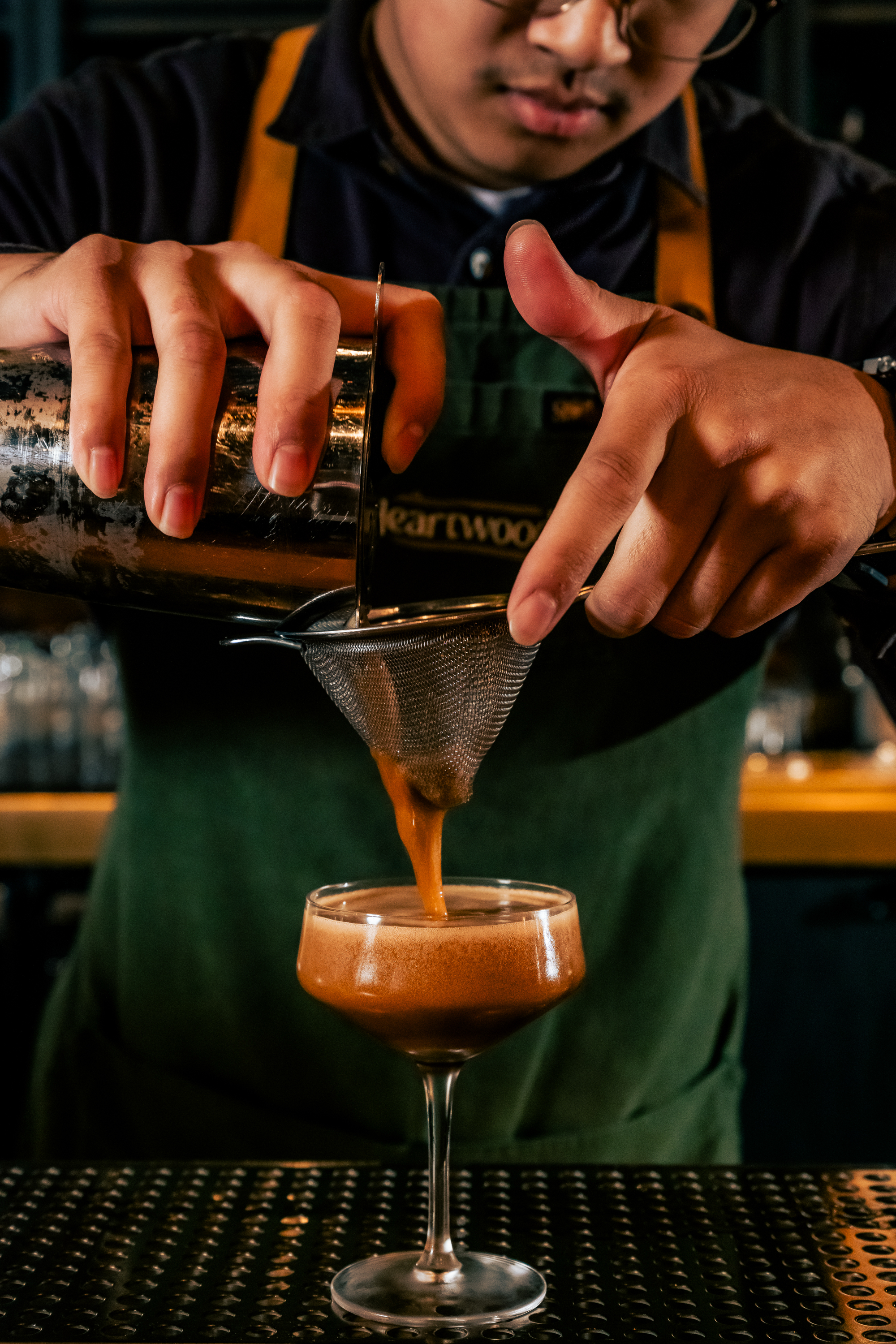 A person is straining a frothy, brown cocktail from a shaker through a fine mesh strainer into a coupe glass, wearing a dark shirt and apron.
