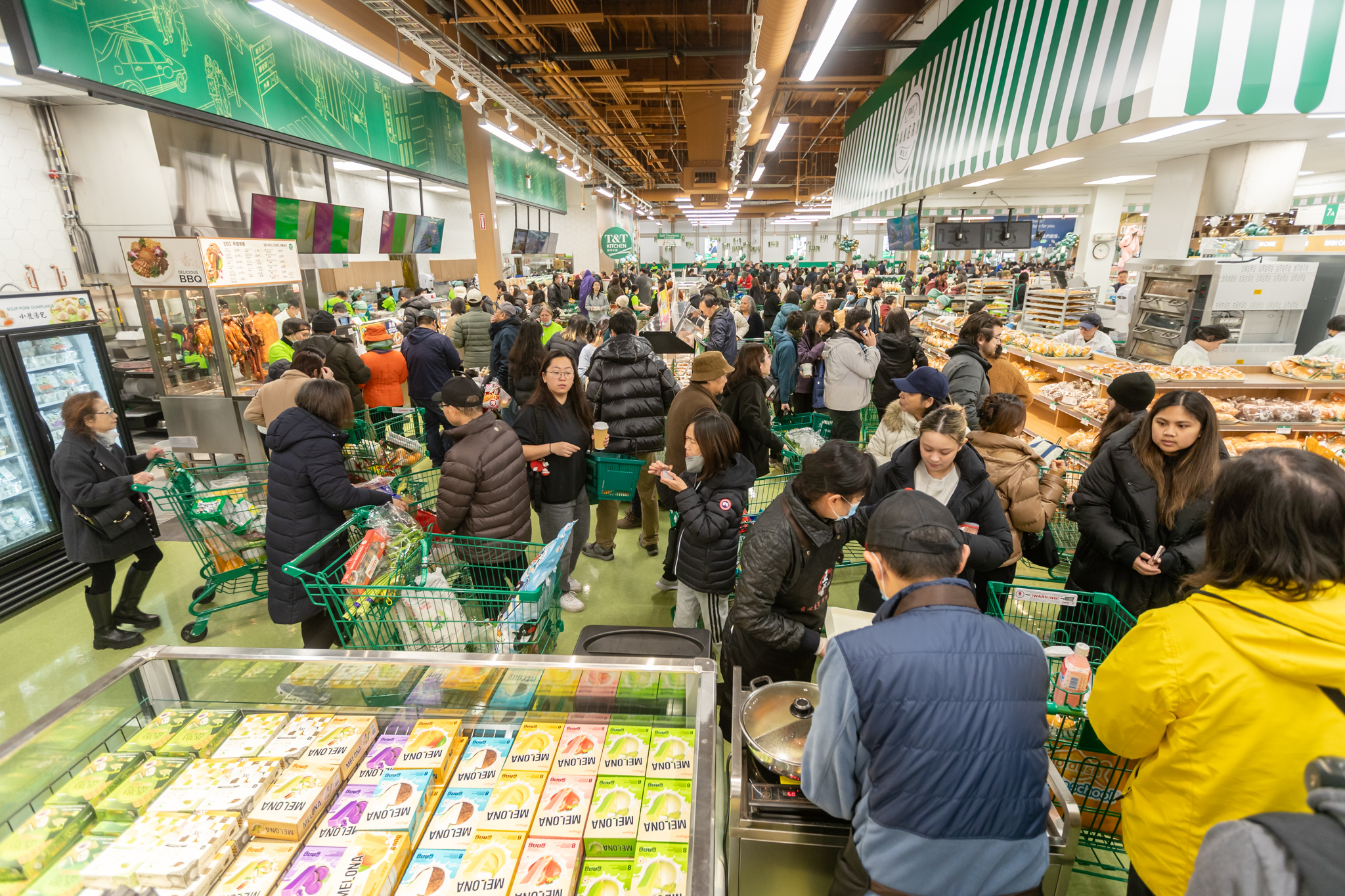 The image shows a crowded supermarket with many people shopping and filling carts. Shelves are stocked with various products, and the environment is busy and bustling.
