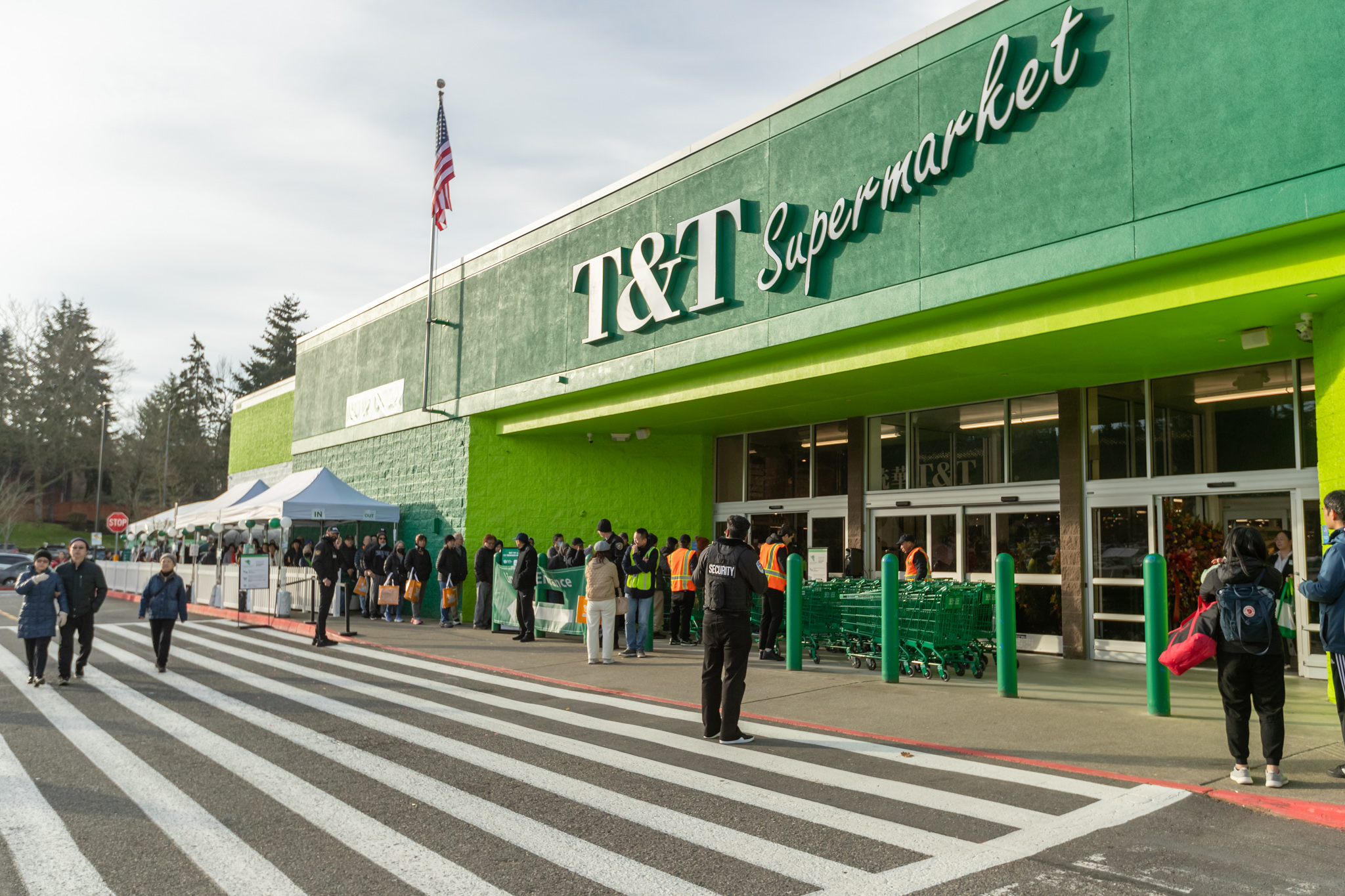 The image shows a crowd outside a T&amp;T Supermarket with a bright green facade. People are lining up under tents, and staff in orange vests manage the entrance.