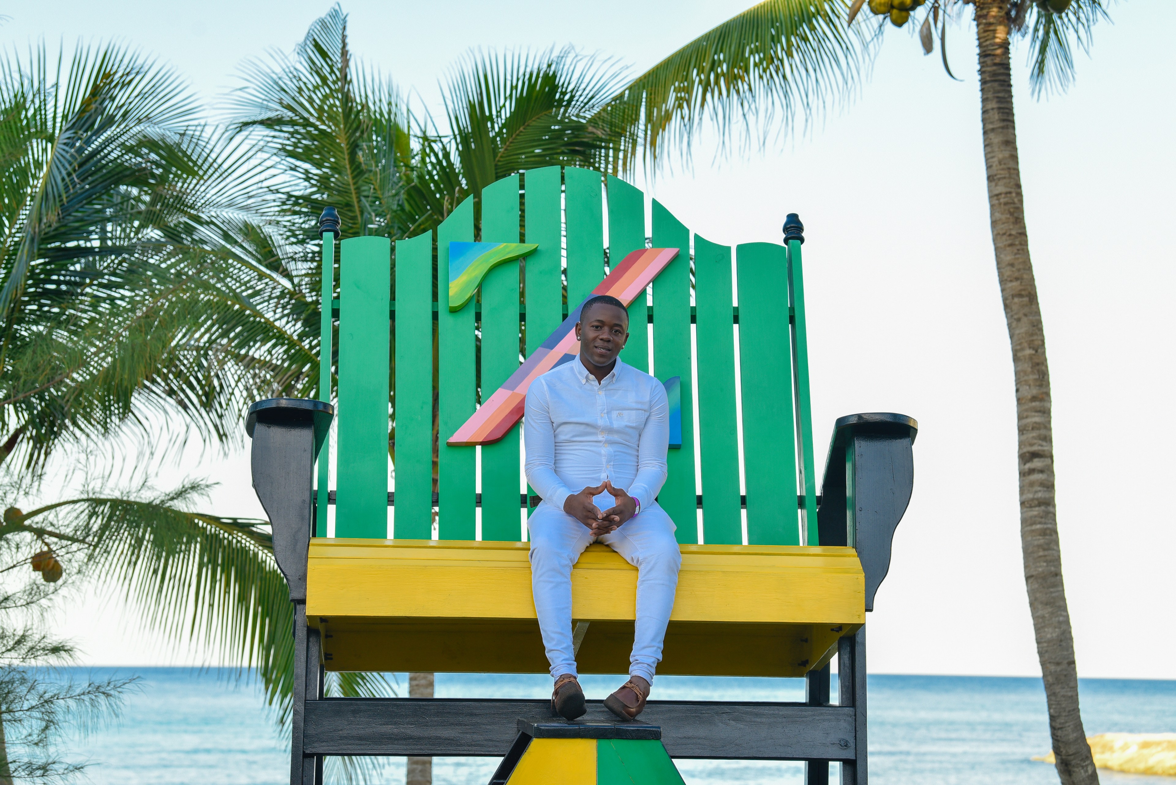 A person in all white sits on a large, colorful chair by the ocean. Palm trees are in the background, and the sea is visible behind them.