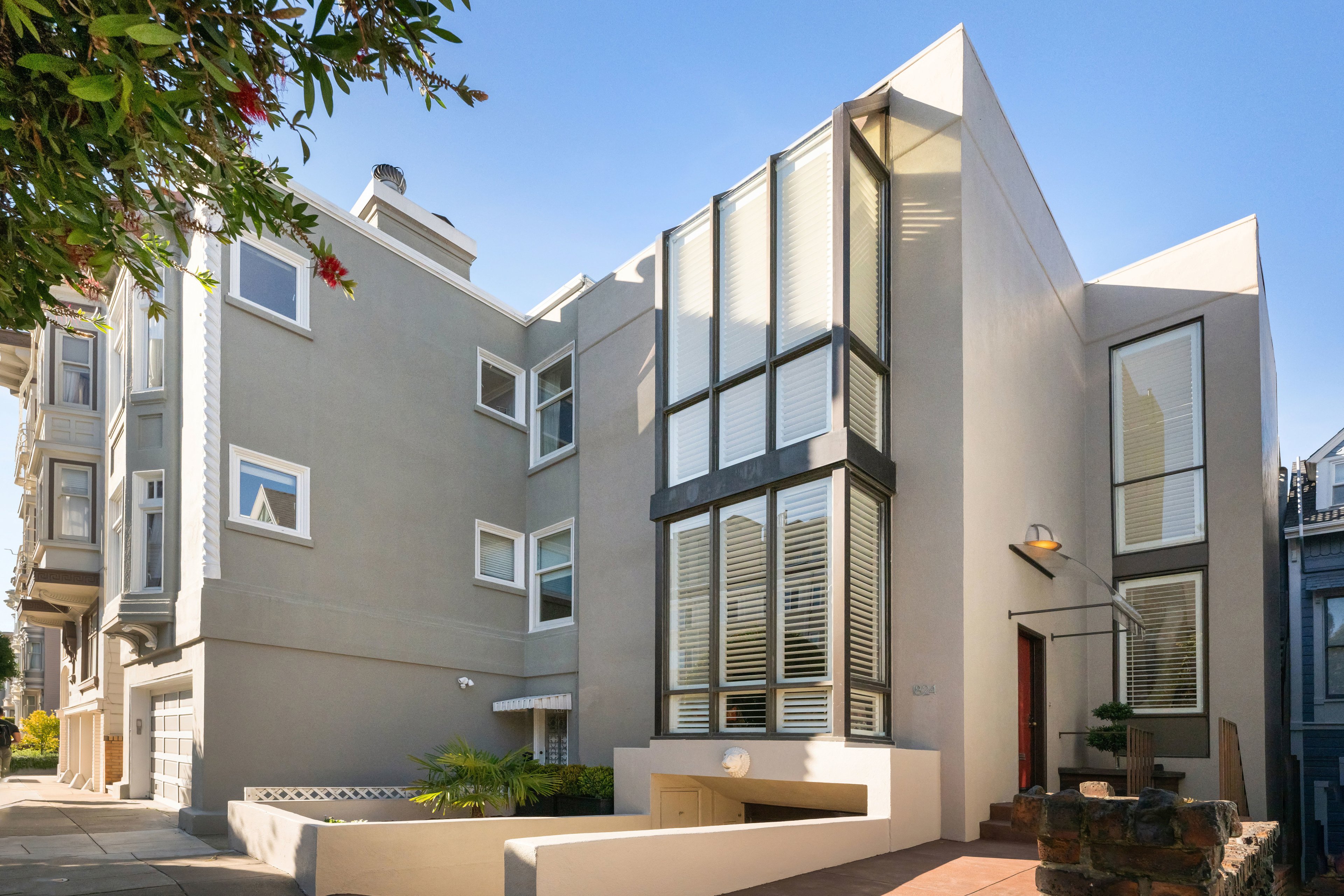The image shows a modern, angular house with large vertical windows and gray walls. It has a small patio area with plants and is adjacent to traditional buildings.