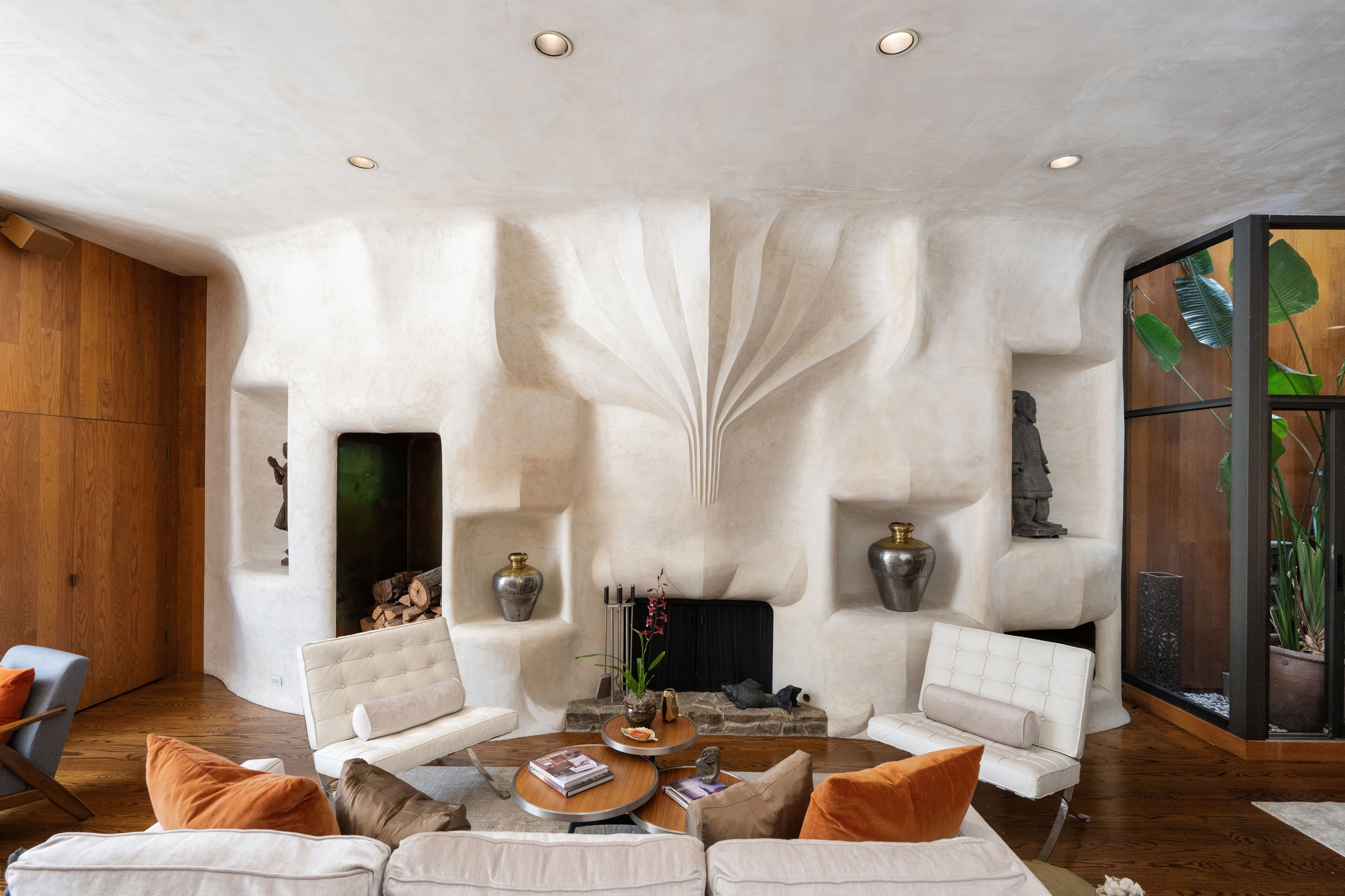 This living room features a sculptural white wall with built-in shelves, pottery, modern chairs, a wooden coffee table, and green plants by a large window.