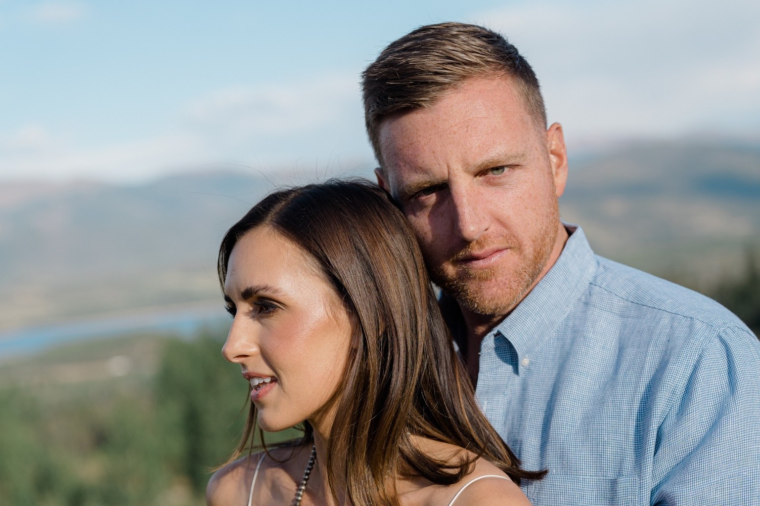 A couple stands embracing outdoors. The woman wears a white dress with turquoise jewelry, while the man is in a blue shirt. The background is a scenic landscape.