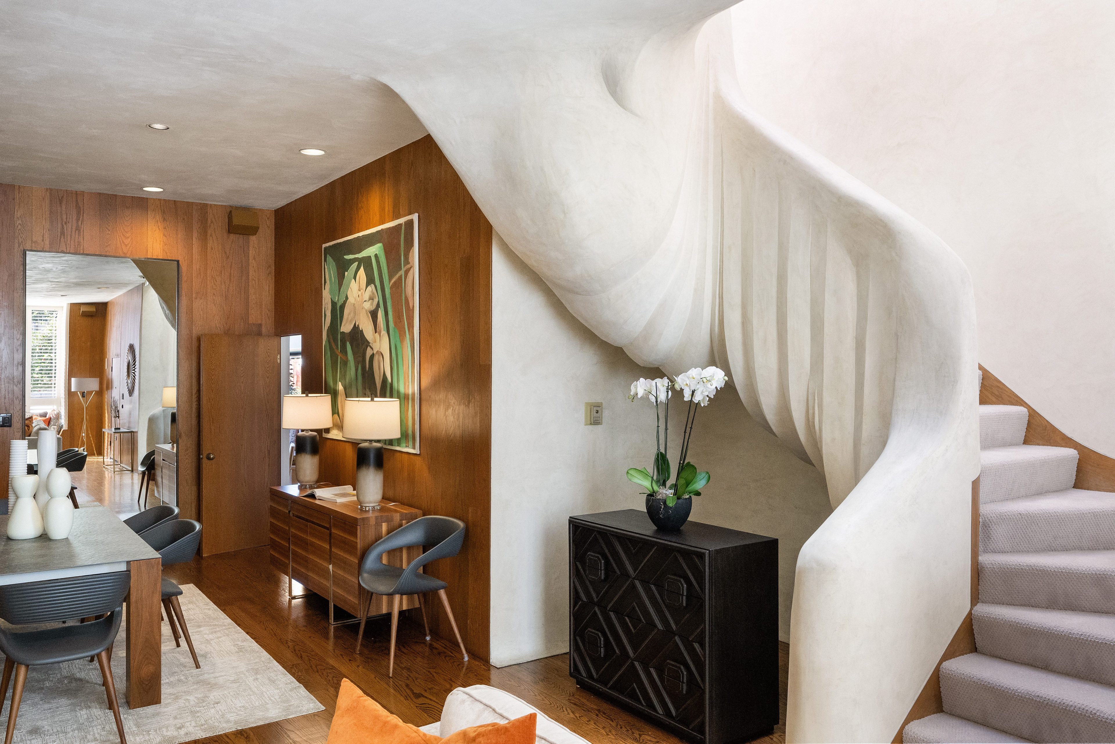 The image shows a modern interior with a sculptural curved staircase, wood-paneled walls, a dining area, and decor including a vase of white flowers on a dark cabinet.