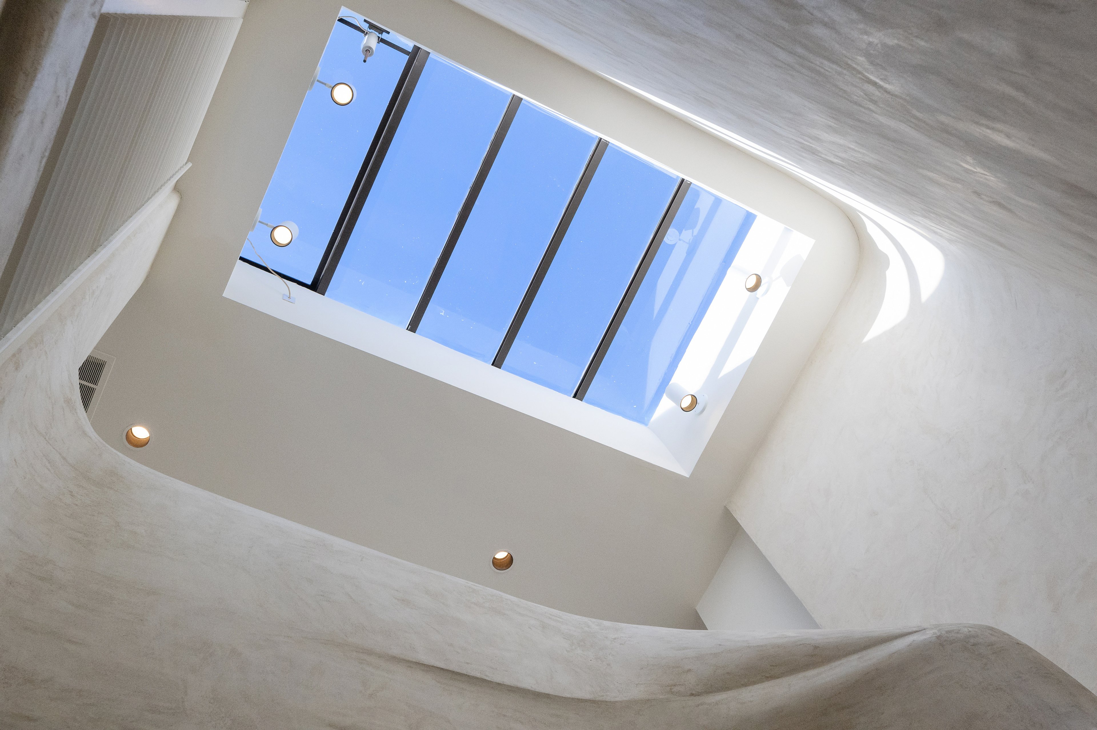 A view looking up at a rectangular skylight with a clear blue sky, surrounded by smooth white walls and ceiling lights embedded around the opening.