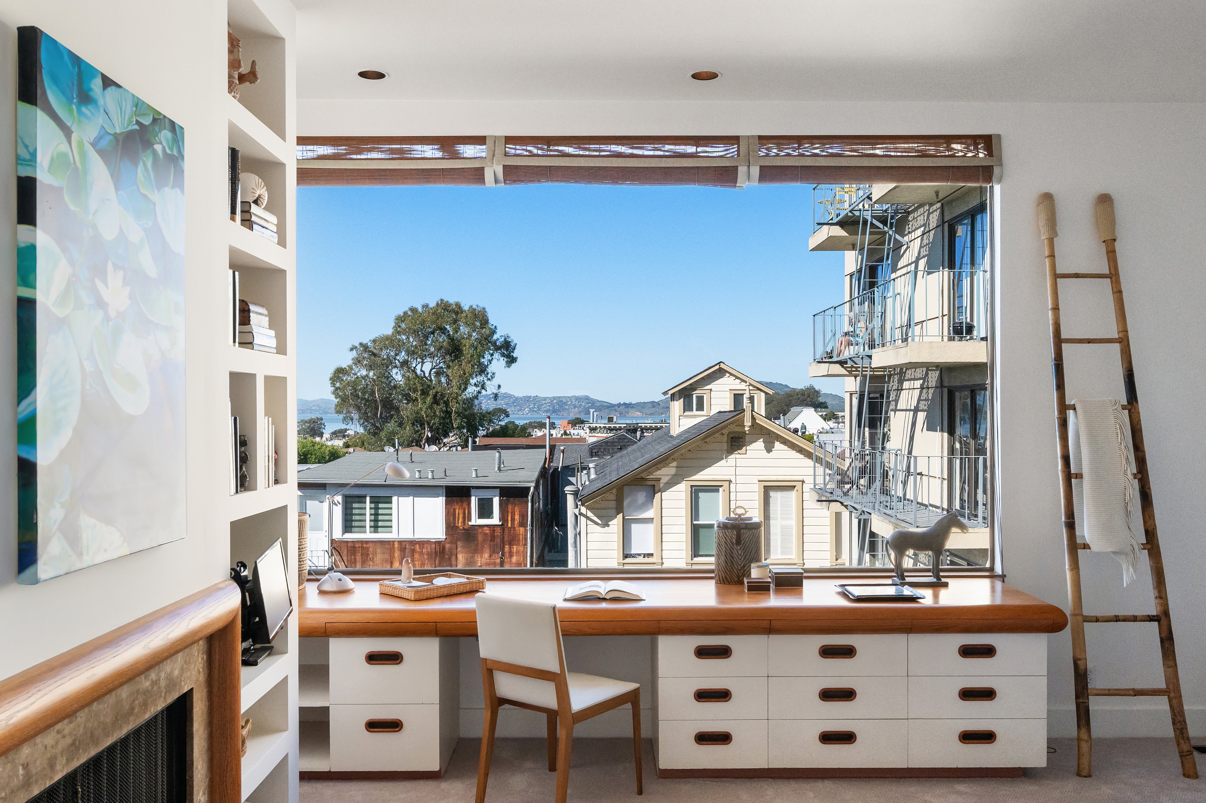 A spacious, bright room with a large desk by a wide window overlooking rooftops and a tree. There's a ladder with a blanket, artwork, and shelves with books.