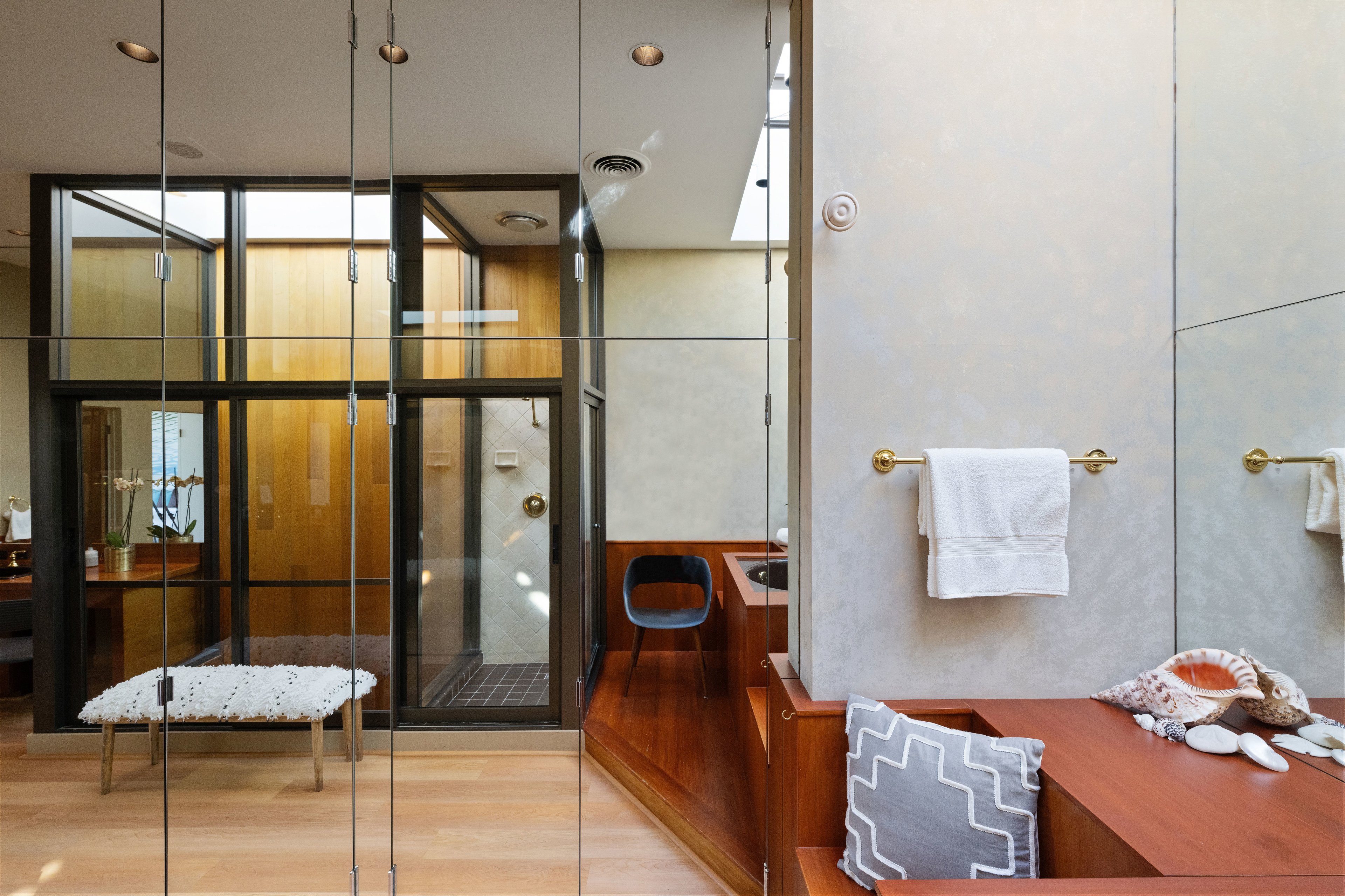 This image shows a modern bathroom with a wooden theme, a glass shower, a bench with a cushion, a towel on a gold holder, and seashells on a wooden surface.