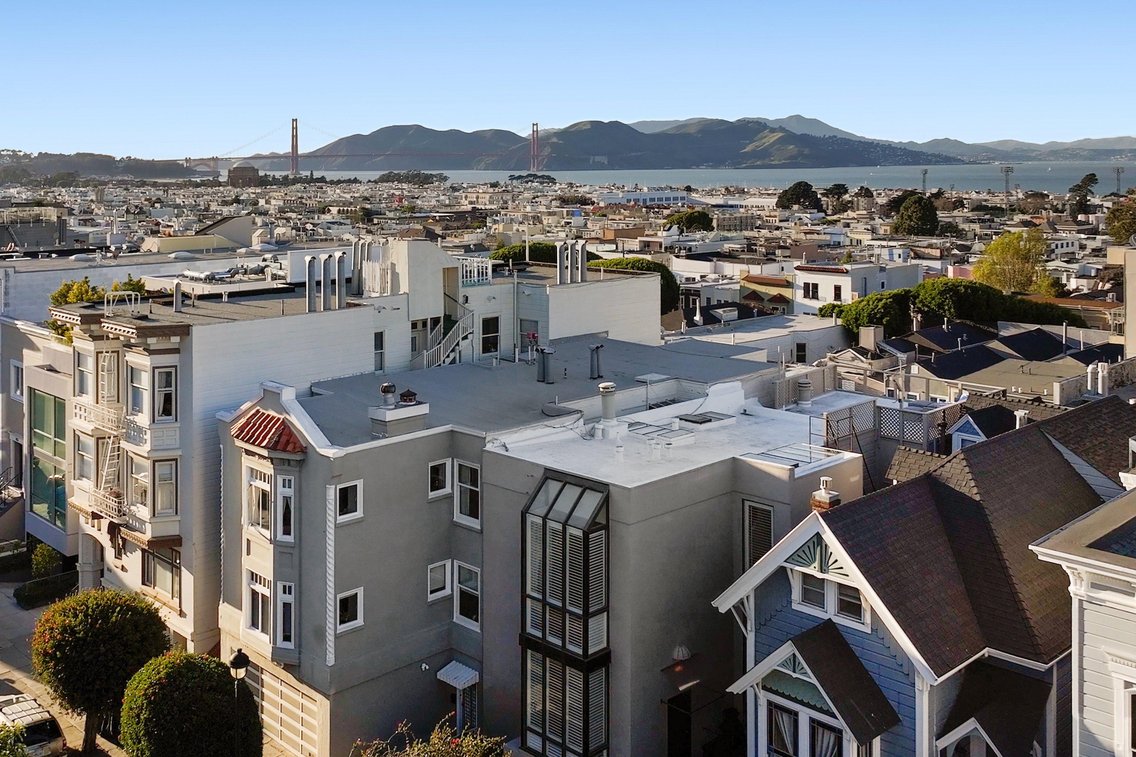 The image shows a cityscape with tightly packed buildings, trees, and the Golden Gate Bridge in the background, under a clear blue sky.