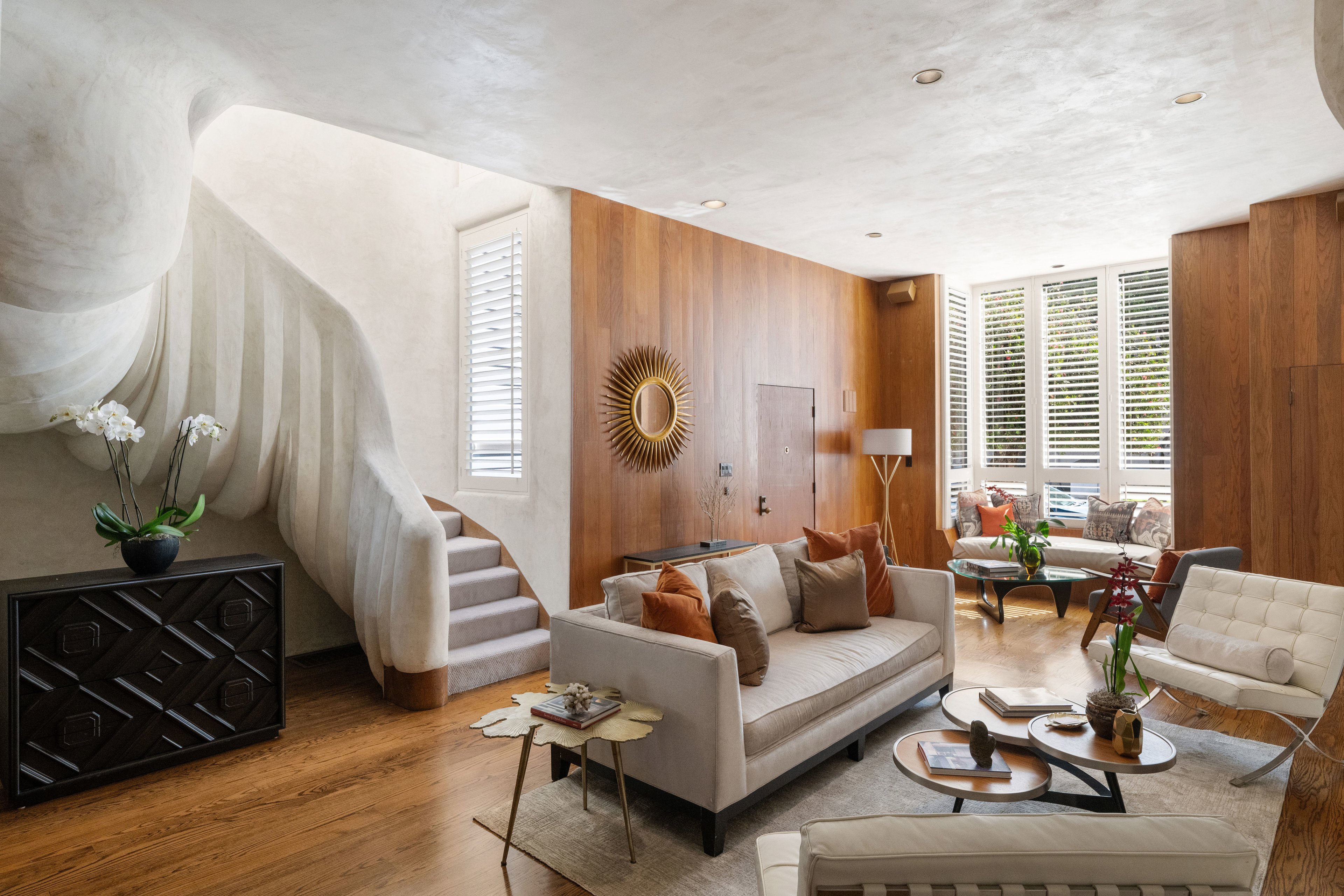 A modern living room features a curved staircase, beige sofa with orange cushions, wood-paneled walls, a sunburst mirror, and a bright window nook.