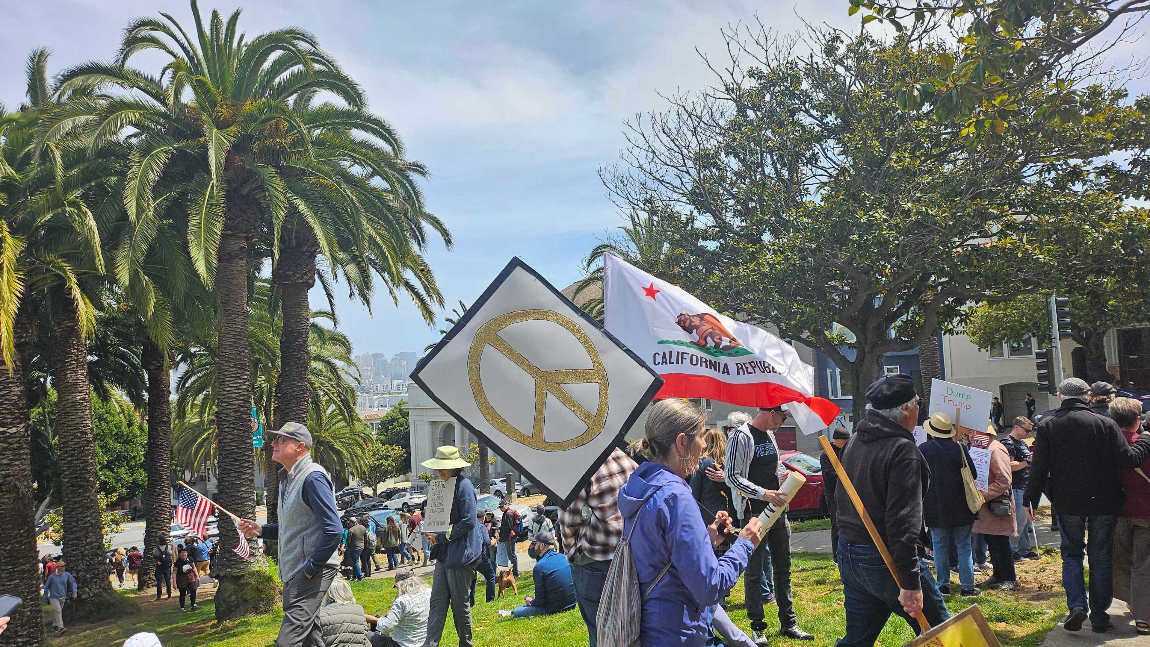 The image shows a park gathering with palm trees and protest signs, including a peace symbol and a California Republic flag. People hold anti-Trump signs.