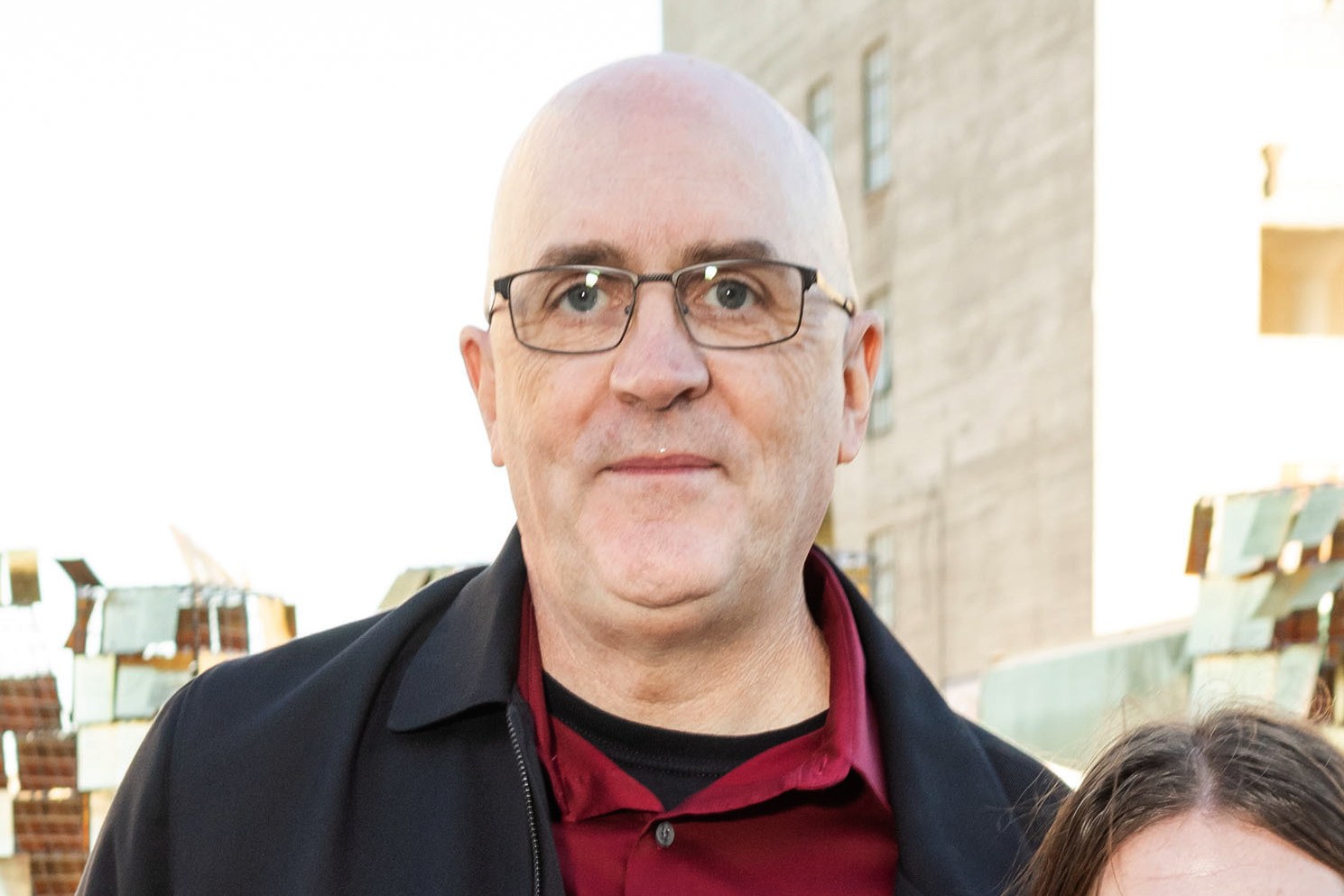 A bald man with glasses is wearing a black jacket over a red shirt, standing outdoors with a building and a metal structure in the background.