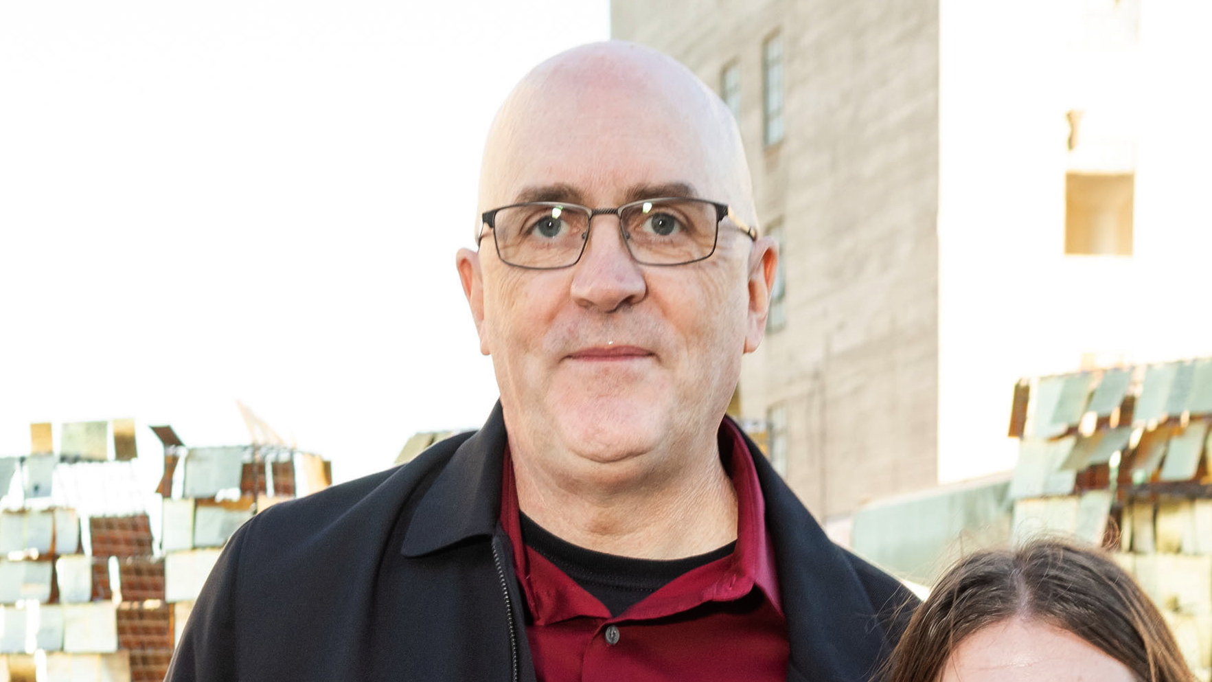 A bald man with glasses is wearing a black jacket over a red shirt, standing outdoors with a building and a metal structure in the background.