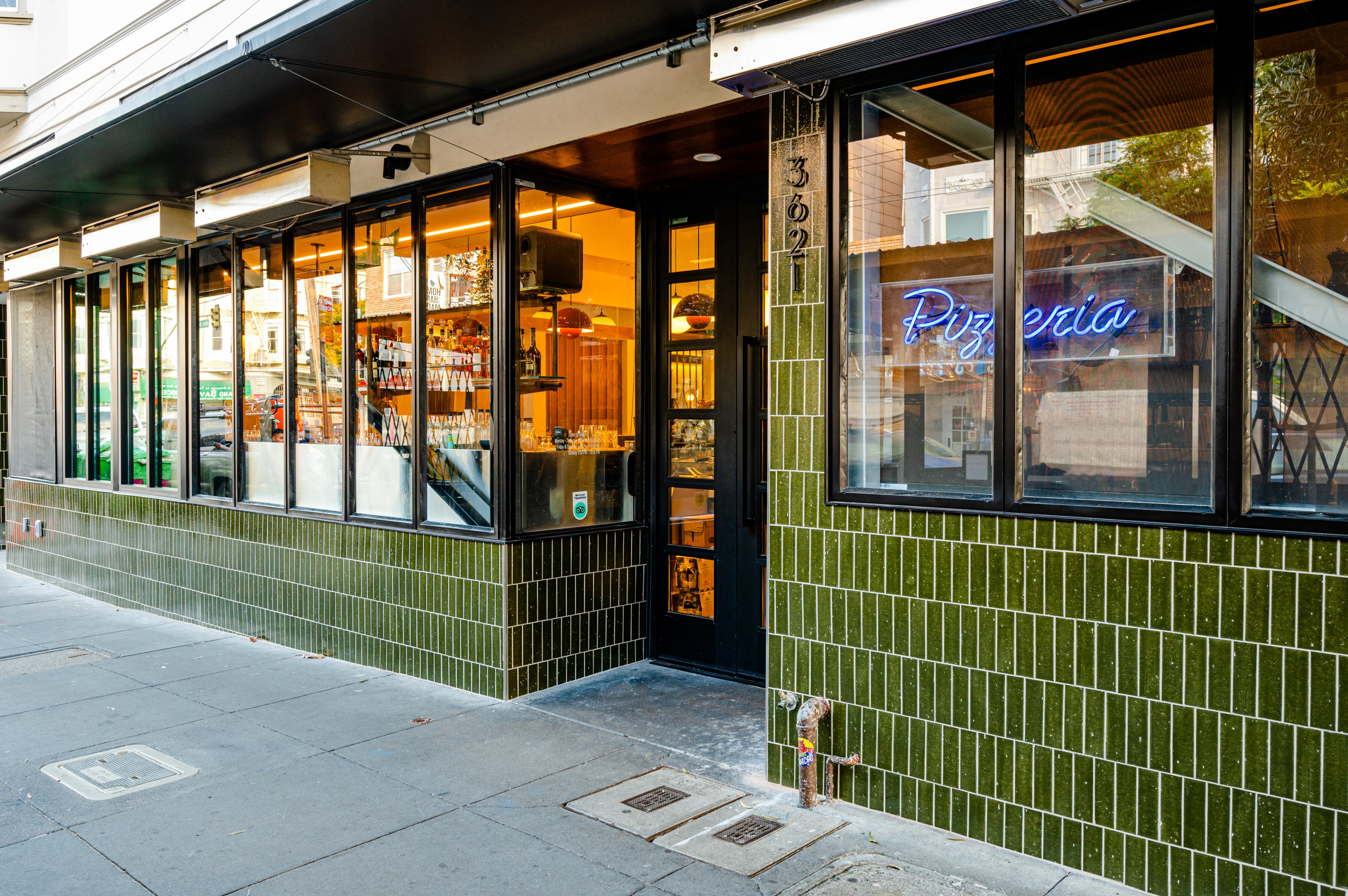 The image shows a pizzeria with a green-tiled exterior, large windows displaying a warm interior, and a neon "Pizzeria" sign. The door is framed by more windows.