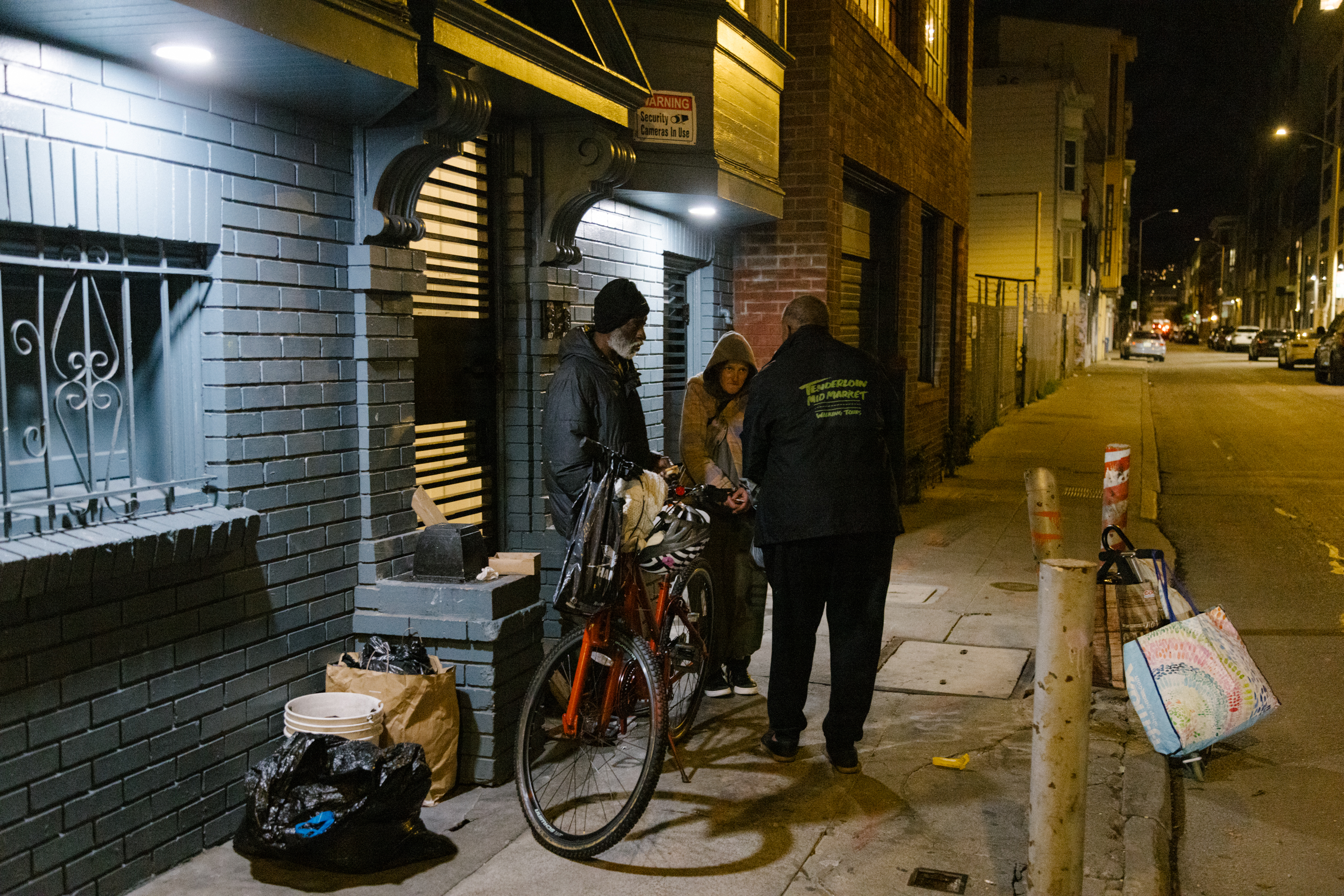 Three people stand on a dimly lit street corner at night. One person has a bicycle. They are next to a blue building with bags and a bucket nearby.