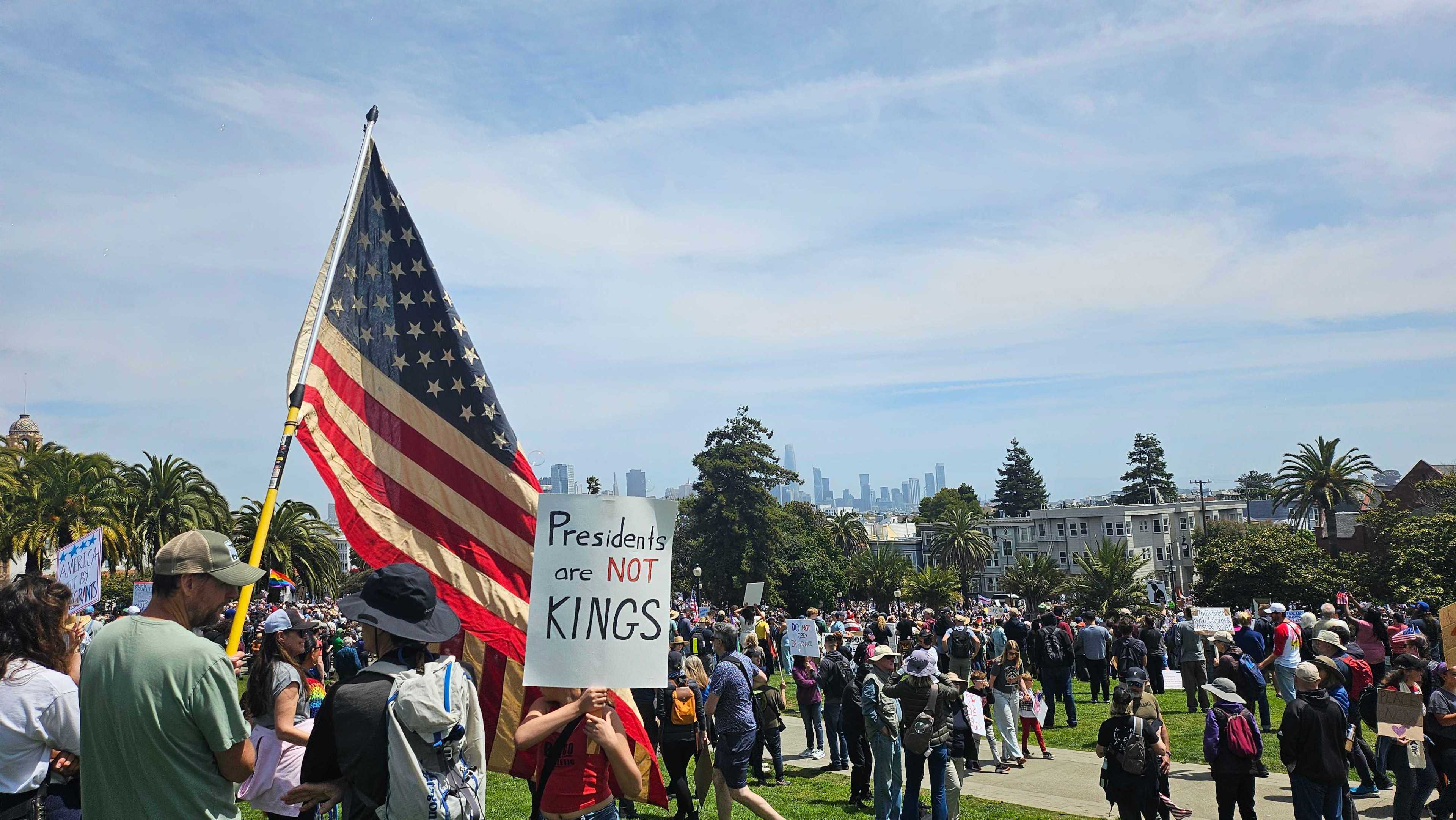A large group of people gather in a park, some holding signs and an American flag. One sign reads "Presidents are not kings." The skyline is visible in the background.