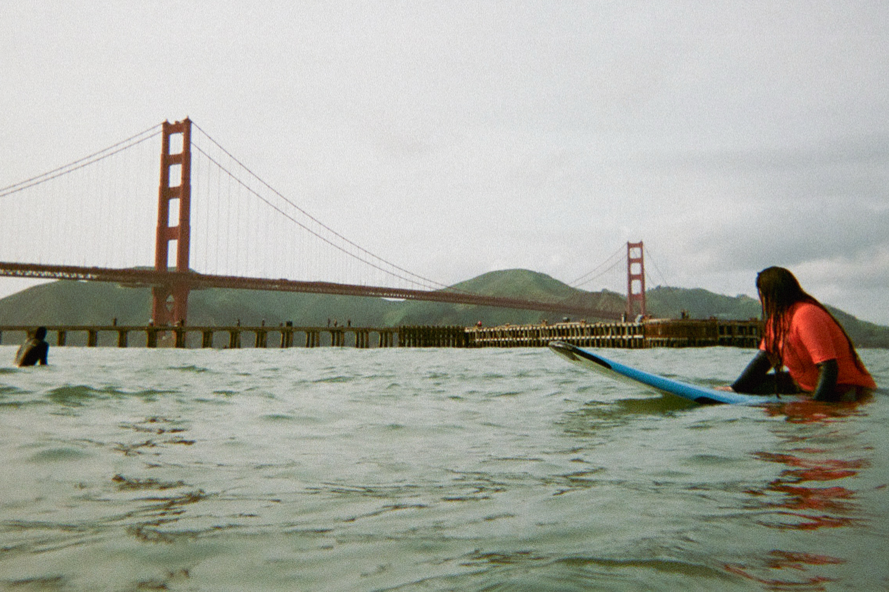 A person on a surfboard sits in the water near the Golden Gate Bridge, with hills in the background and a pier extending out into the water.