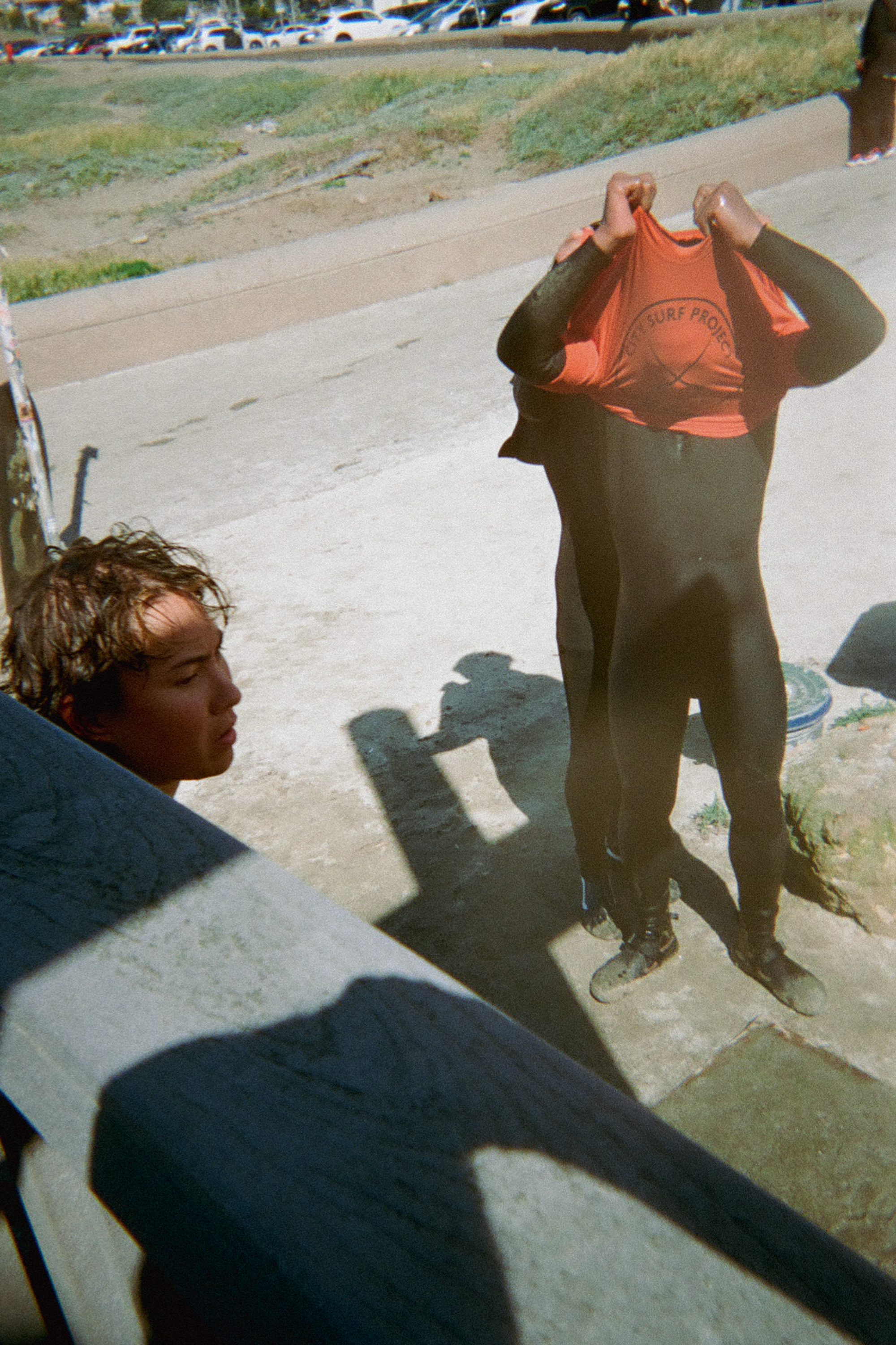 A person wearing a wetsuit pulls a red shirt over their head. Another person stands nearby, while shadows and a grassy area with parked cars are visible.