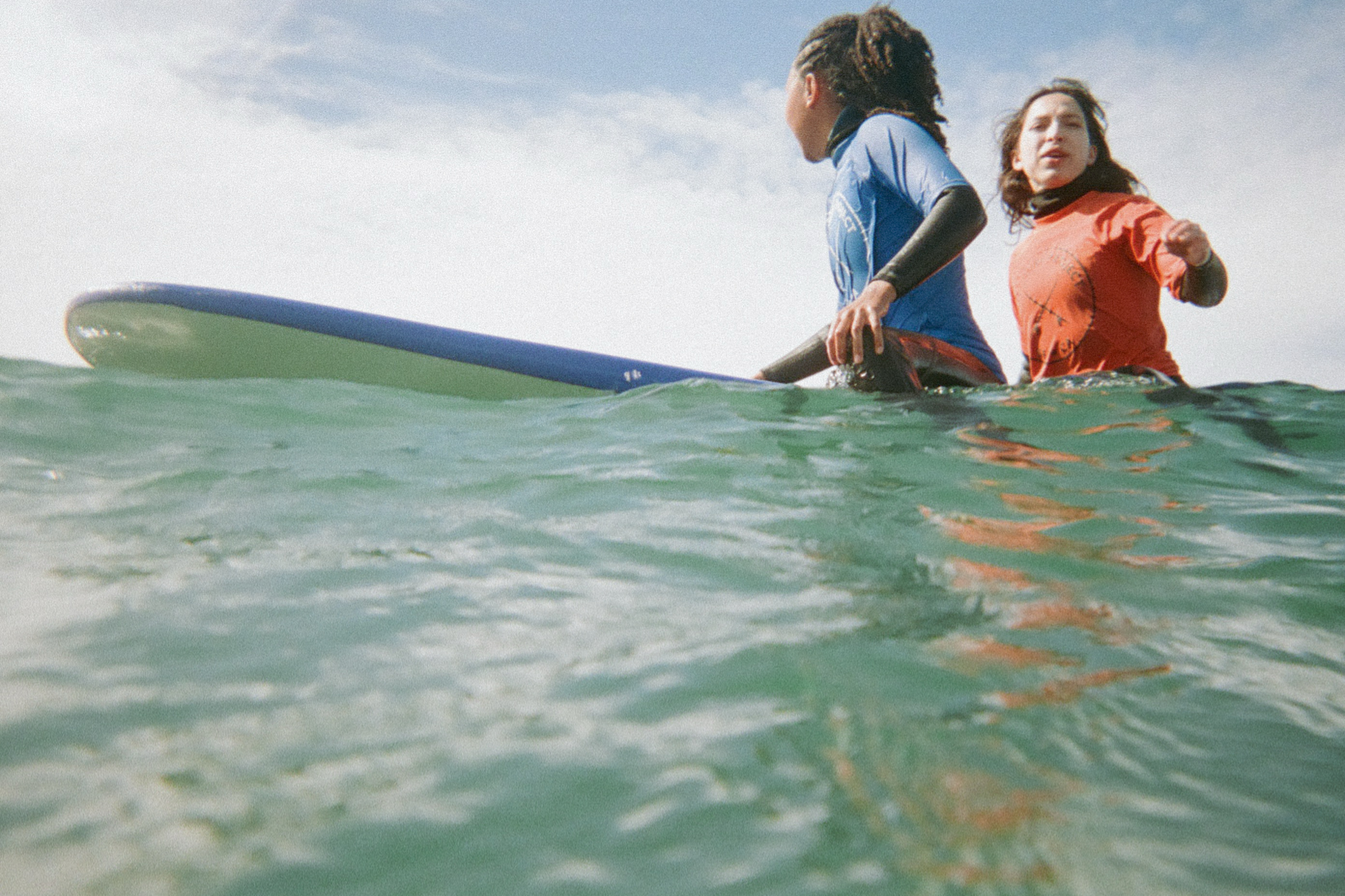Two people in wetsuits sit on a surfboard in the ocean, with one wearing blue and the other red. They are surrounded by greenish water under a cloudy sky.