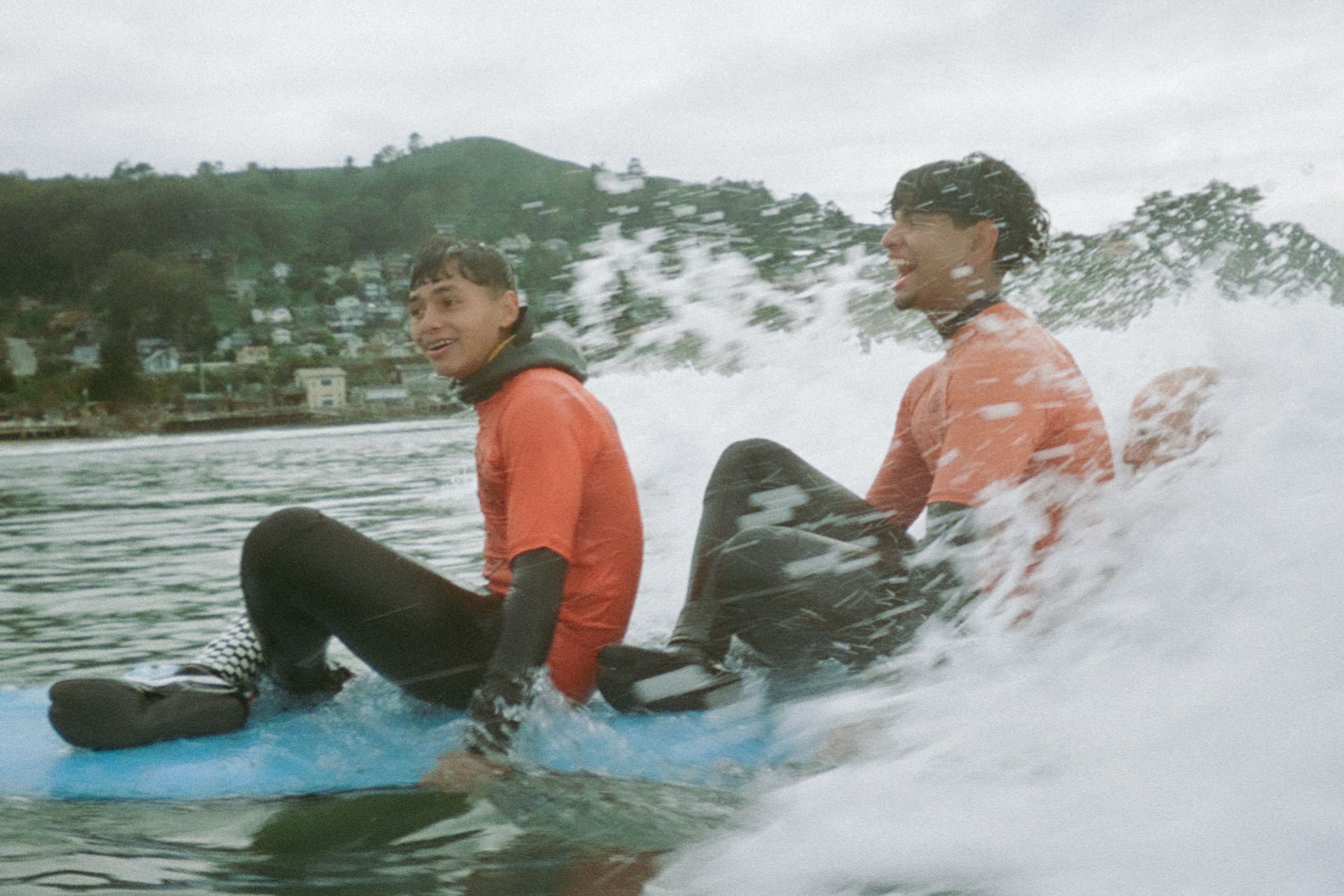 Two people in wetsuits and orange shirts are sitting on a surfboard in the ocean, smiling as a wave splashes around them. Hills and houses are visible in the background.