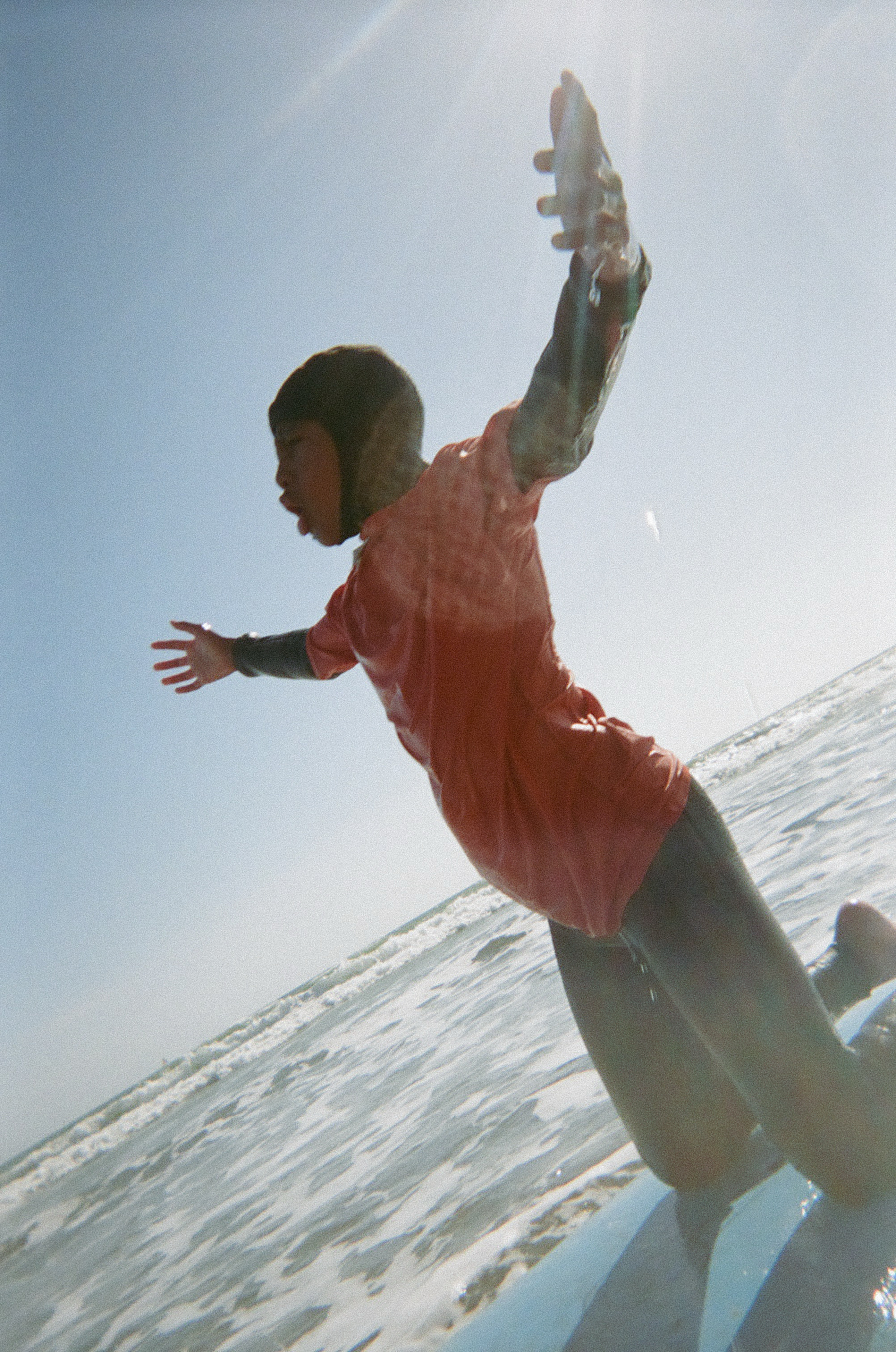 A person in a red shirt and wetsuit kneels on a surfboard in the ocean, with arms outstretched as sunlight beams down from a clear sky.