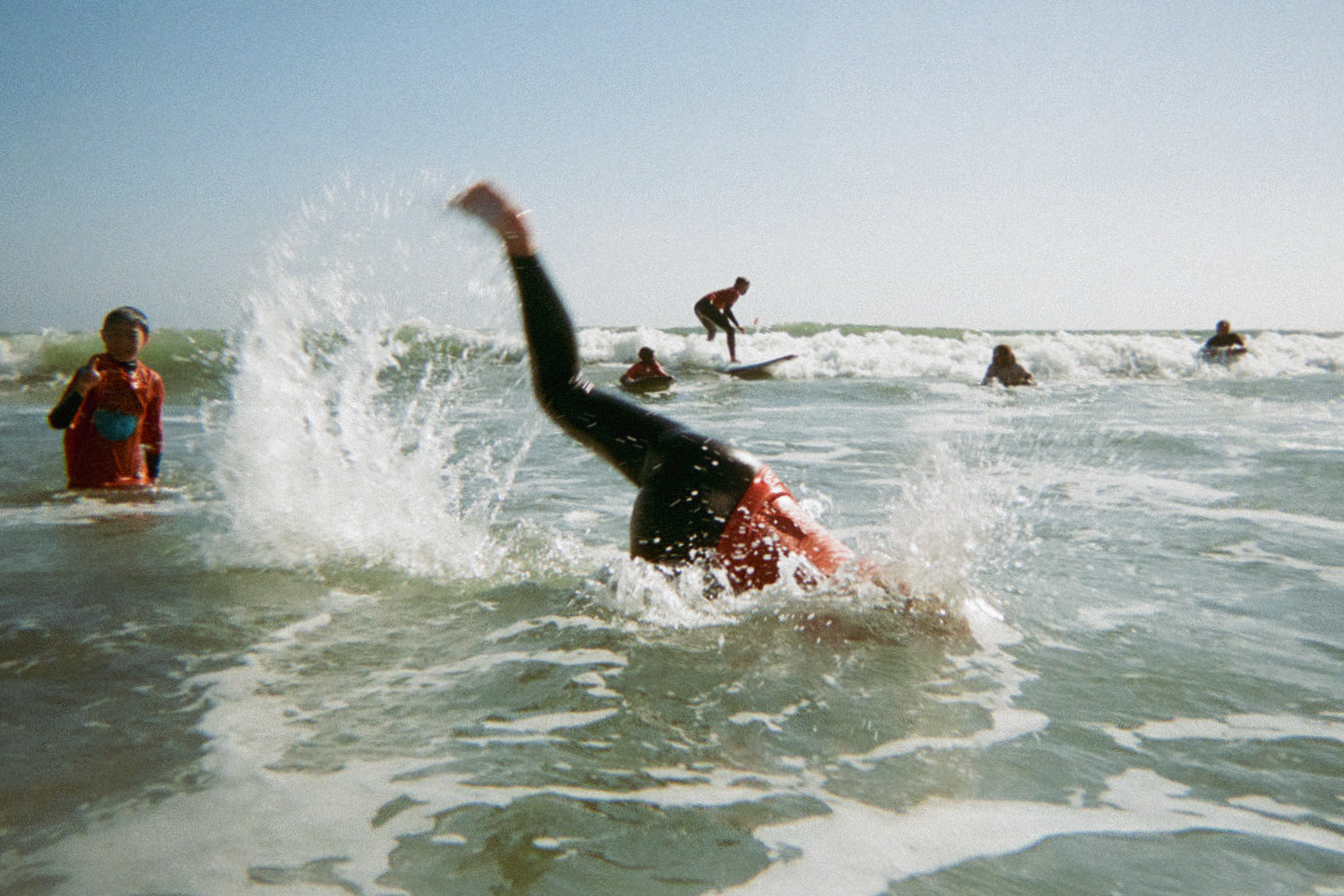 A person in a red shirt falls headfirst into the water, creating a splash. Nearby, others are surfing on small waves under a clear blue sky.