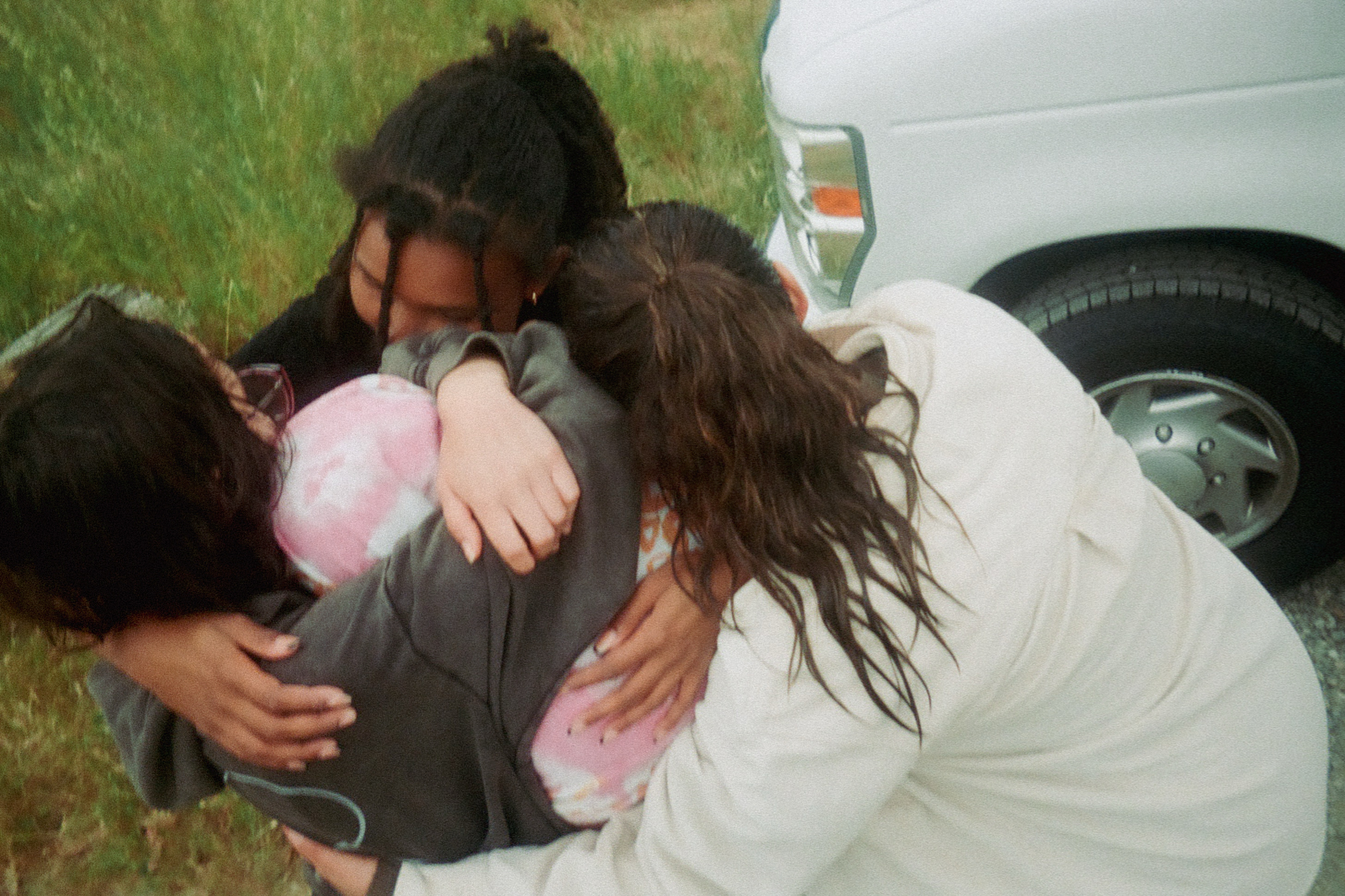 Four people are in a group hug near a white vehicle, with grass in the background. They are wearing casual, comfy clothing, expressing closeness and comfort.