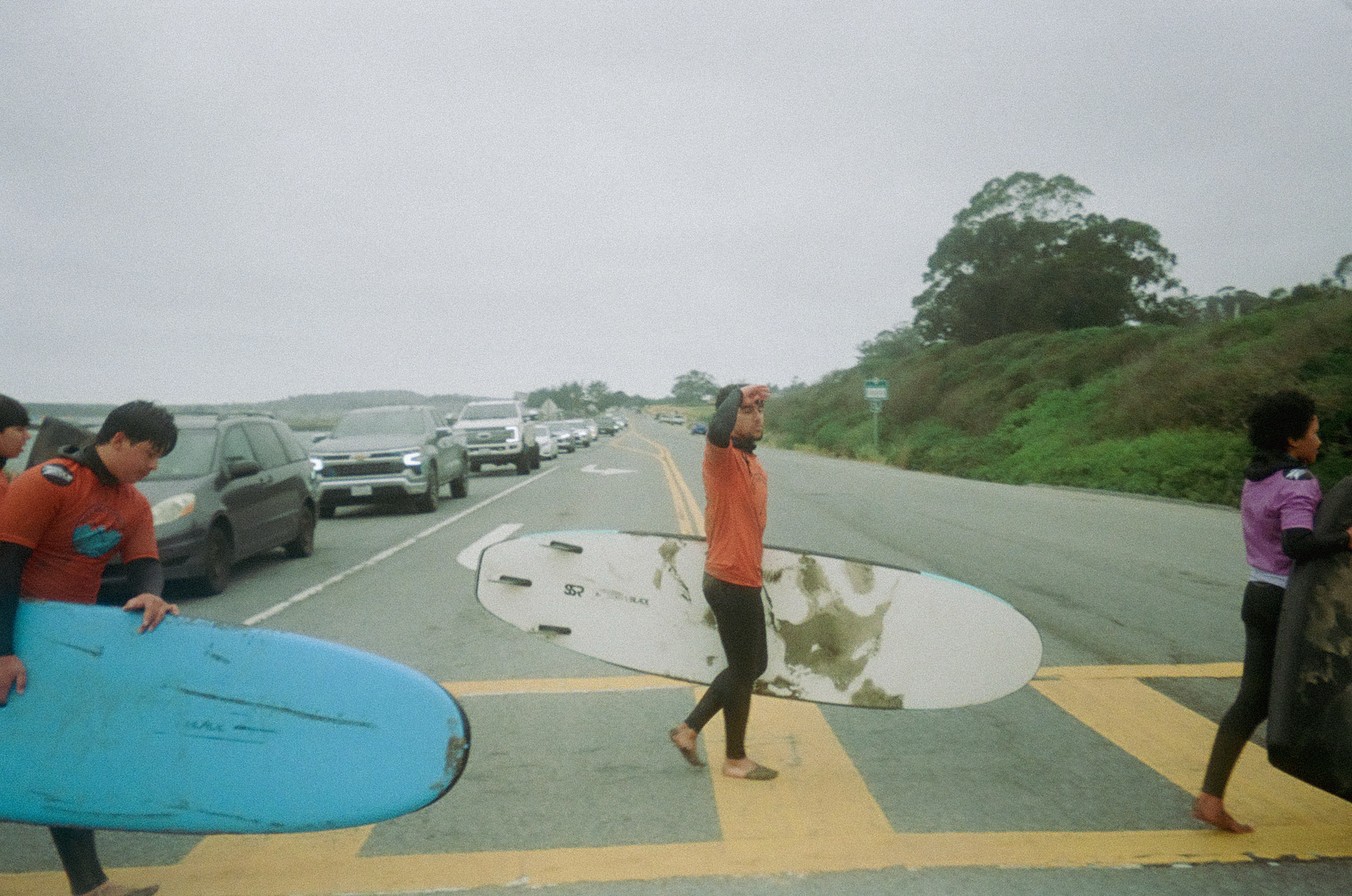 Three people, wearing wetsuits and holding surfboards, cross a road with cars queued nearby. The sky is overcast, and greenery lines the road.