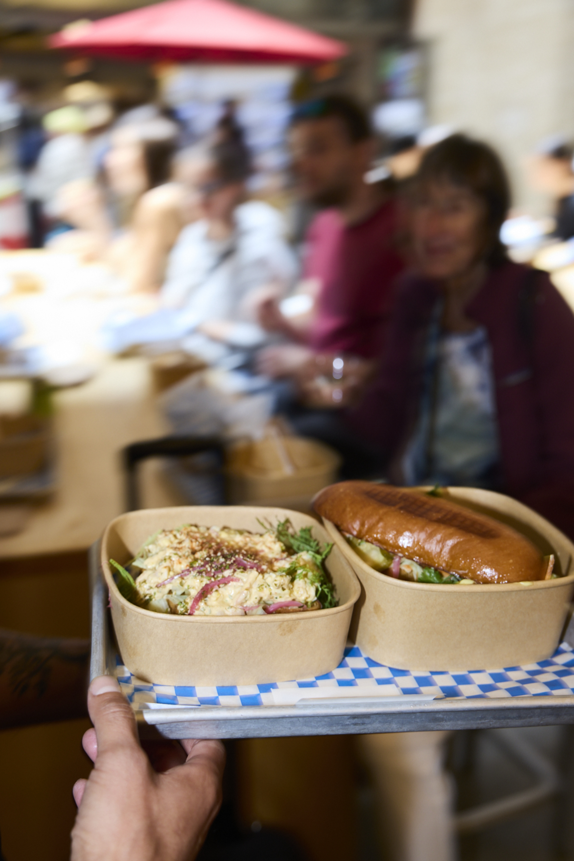 A hand holds a tray with a salad and sandwich in disposable containers. The background shows blurred people seated at a table, with a red umbrella overhead.