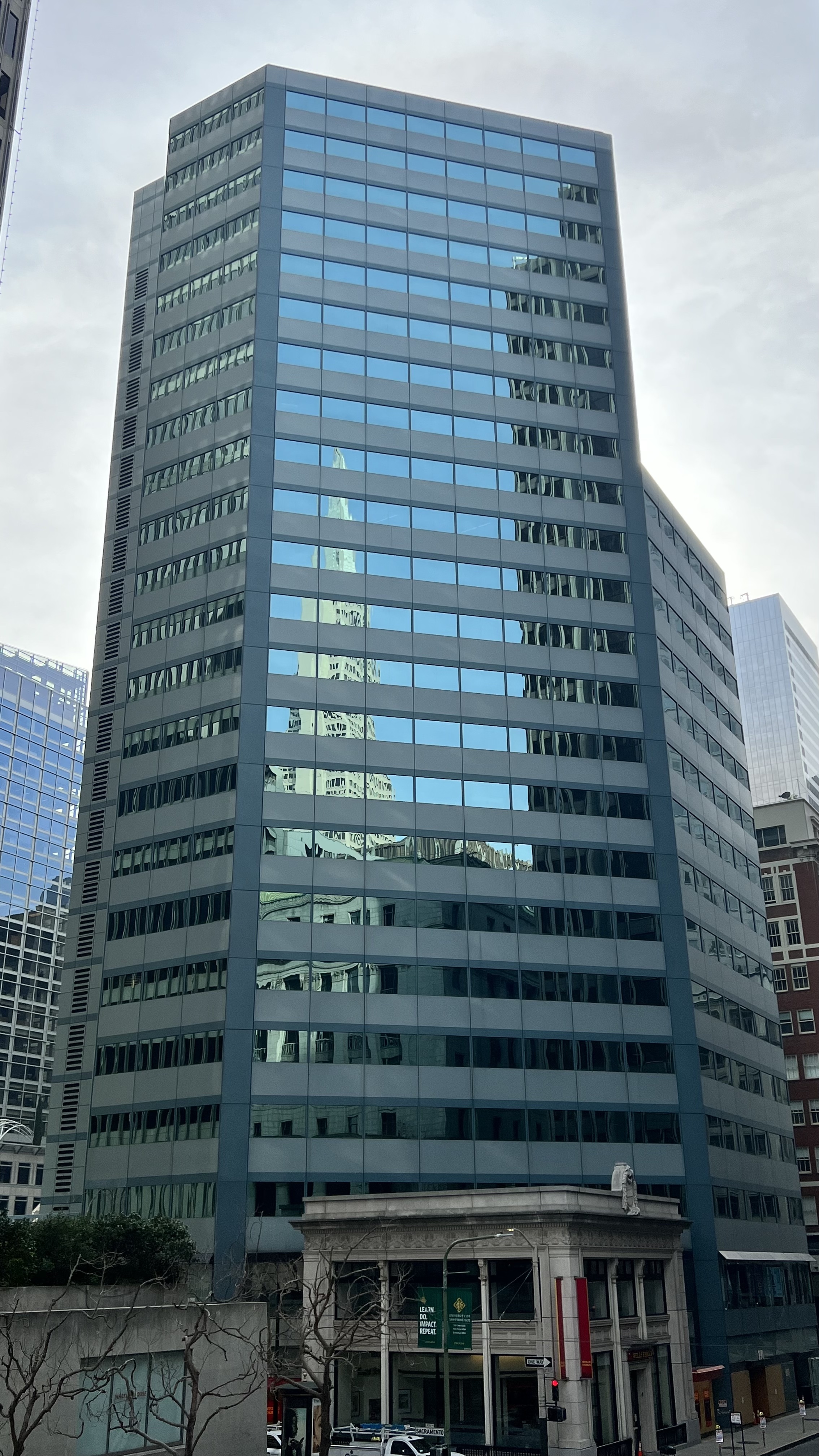 A tall building with reflective blue windows shows a distorted reflection of another skyscraper. A section of a shorter building with columns is at the base.