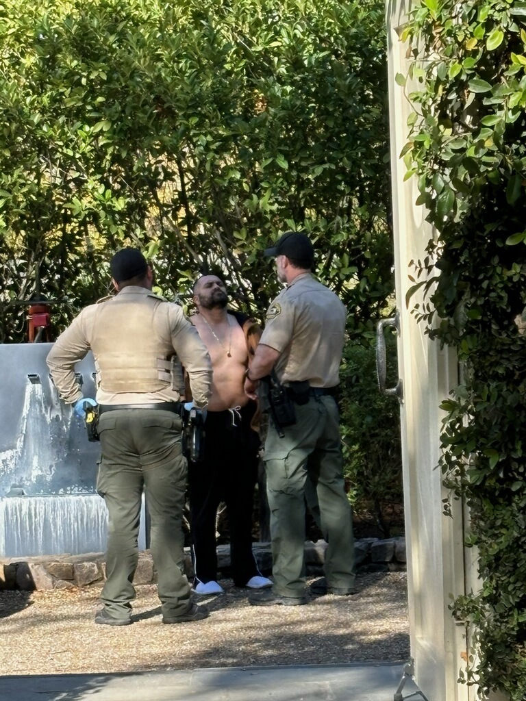 Two officers stand facing a shirtless man outdoors, near a fountain and greenery. They appear to be in conversation, surrounded by lush plants.
