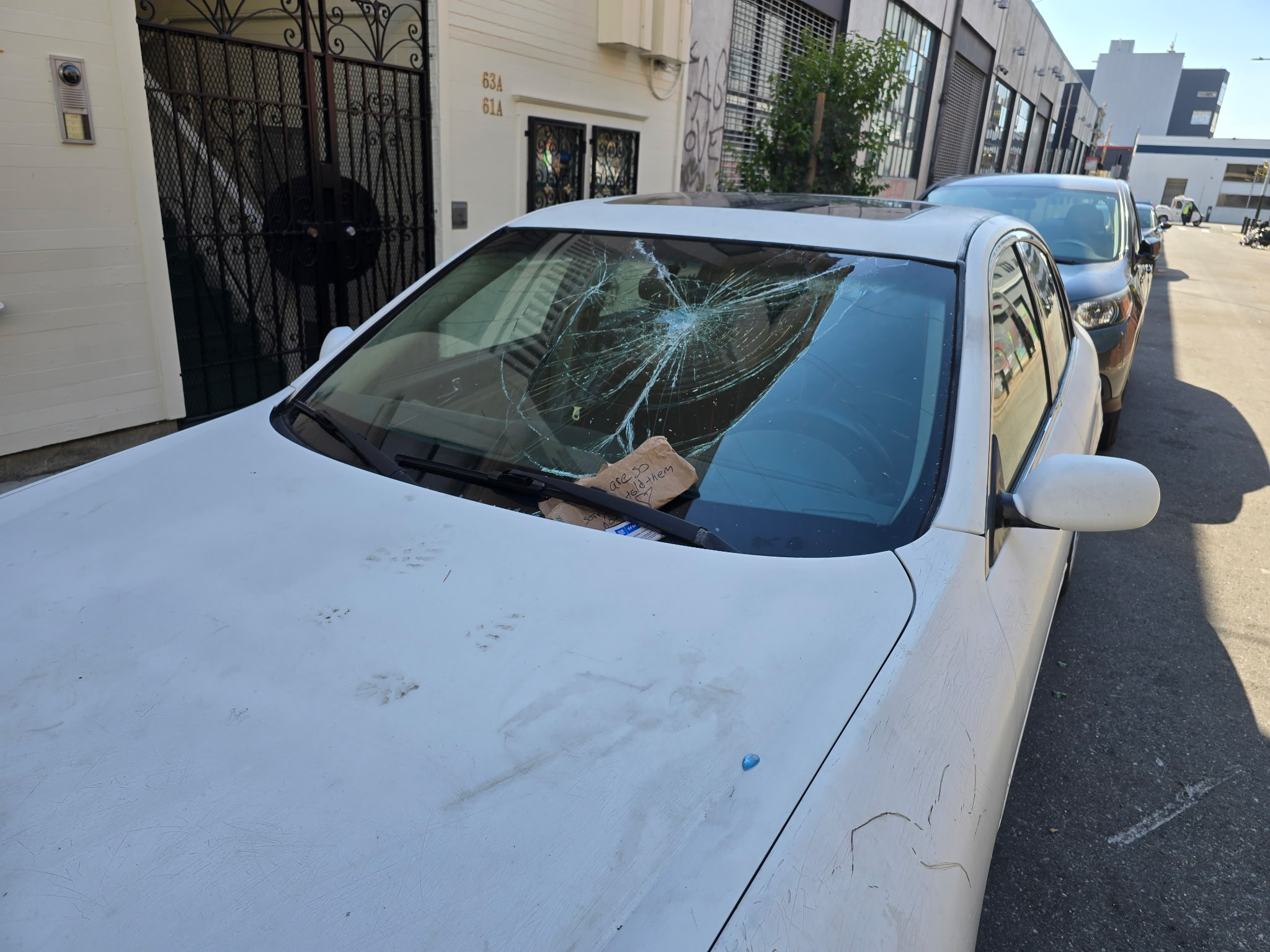 A white car is parked on the street with a shattered windshield and visible scratches. There's a note under the wiper, and the car is near two other parked vehicles.