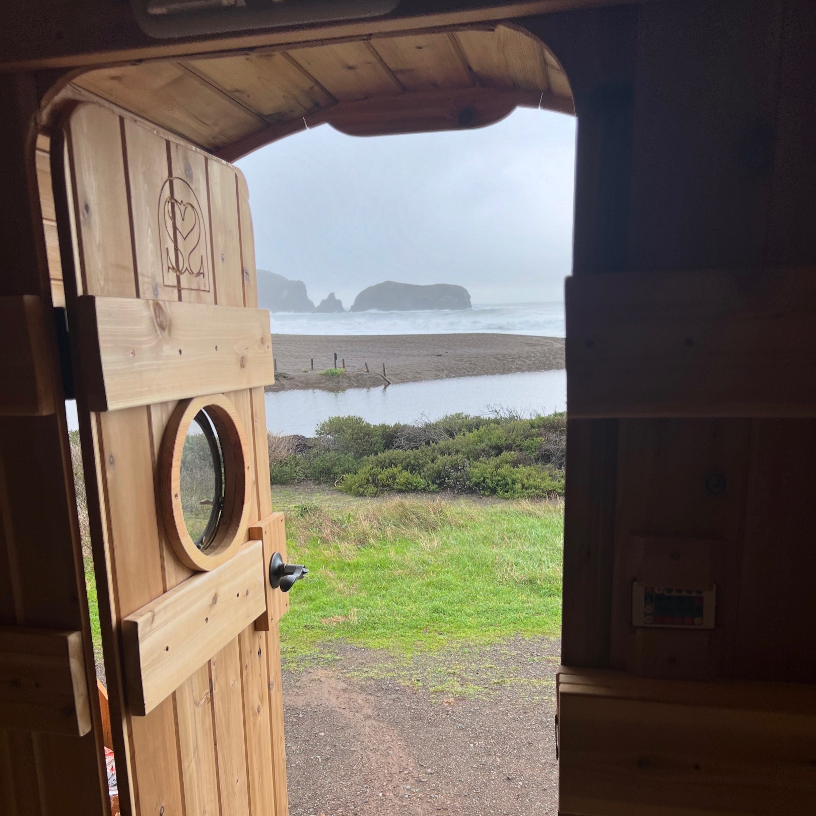 A wooden door with a round window opens to a coastal view. There are rocky formations in the sea, sandy beach, grassy area, and overcast sky.