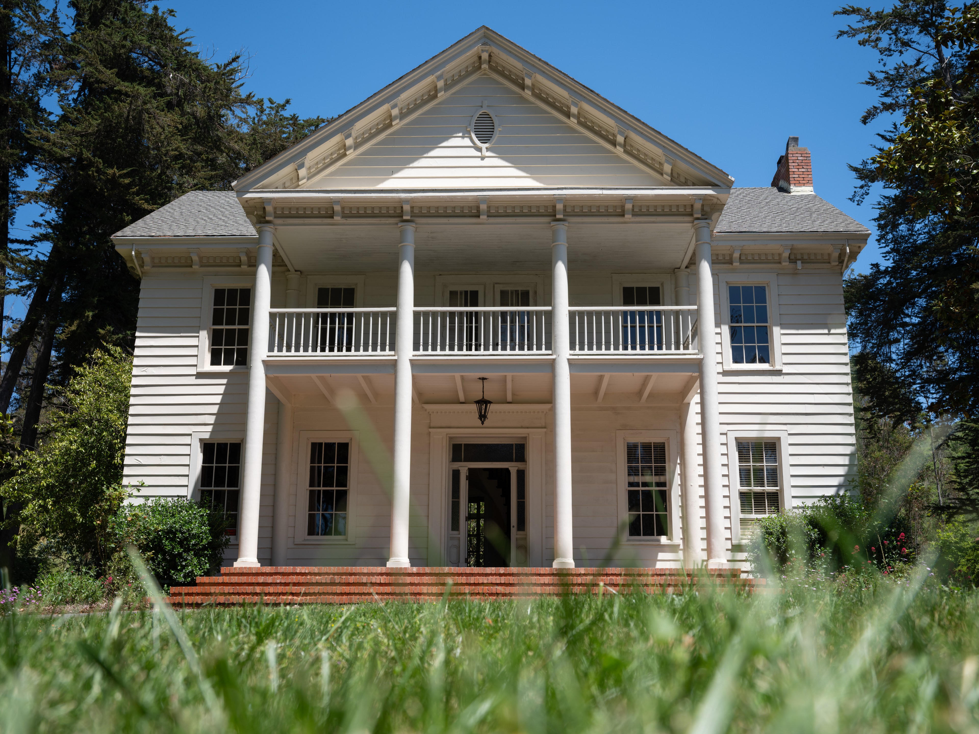 A two-story white house with tall pillars stands against a clear blue sky, surrounded by green trees and shrubs, with a brick step entrance in the foreground.