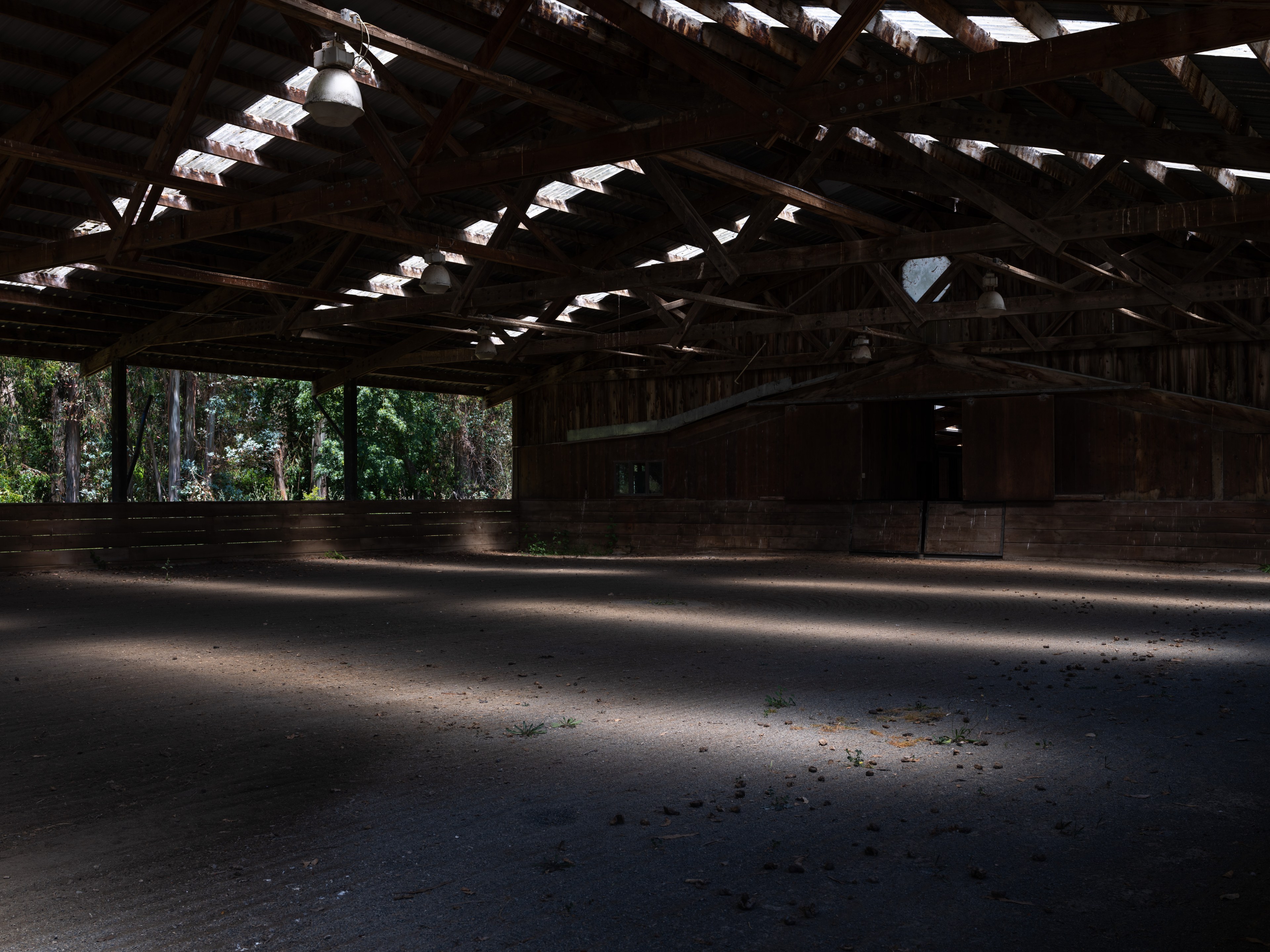 The image shows a dimly lit, empty wooden structure with a high ceiling and beams. Sunlight streams in through gaps, creating patterns on the dusty floor.