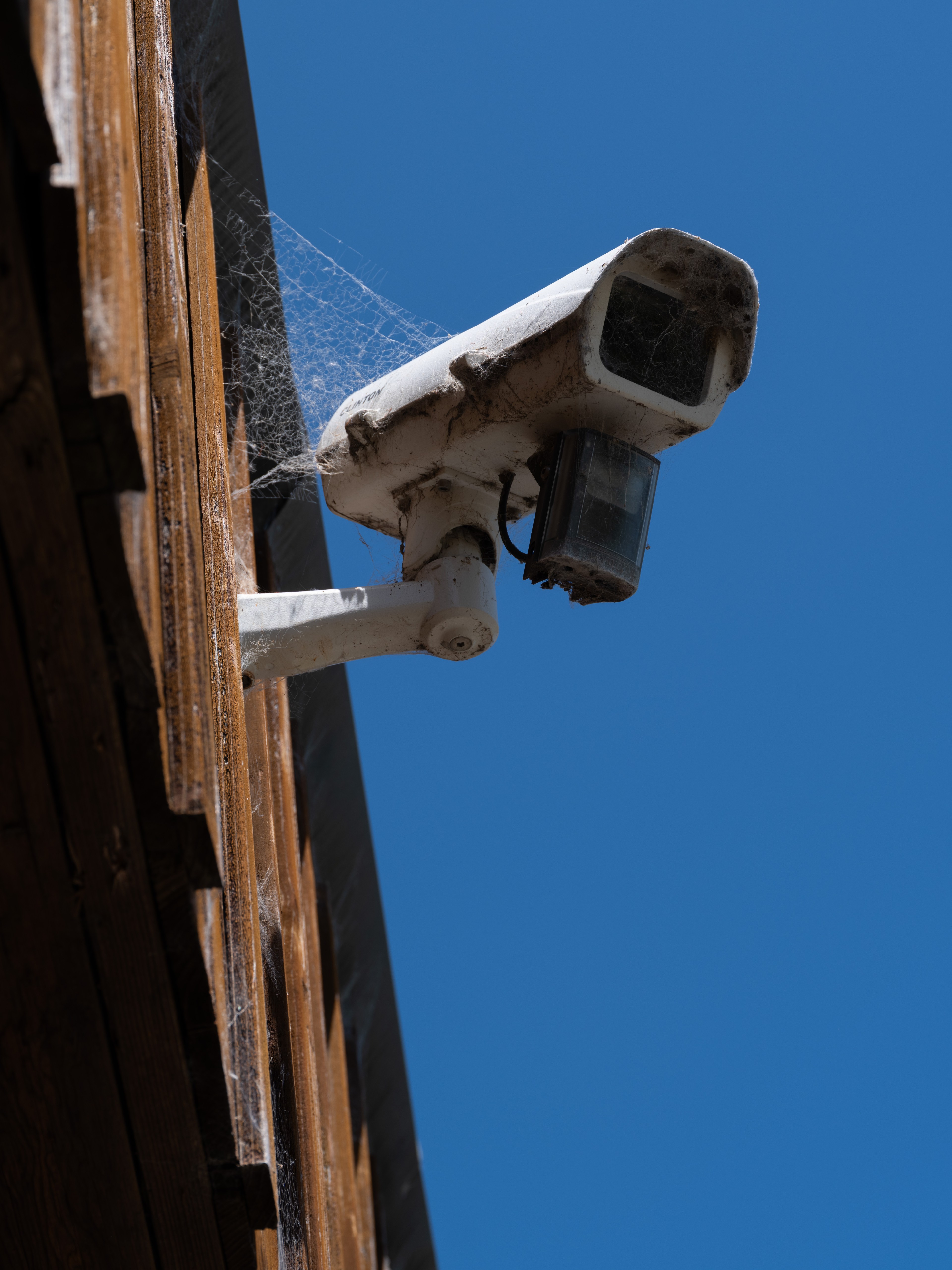 A weathered security camera with cobwebs is mounted on a wooden wall against a clear blue sky.