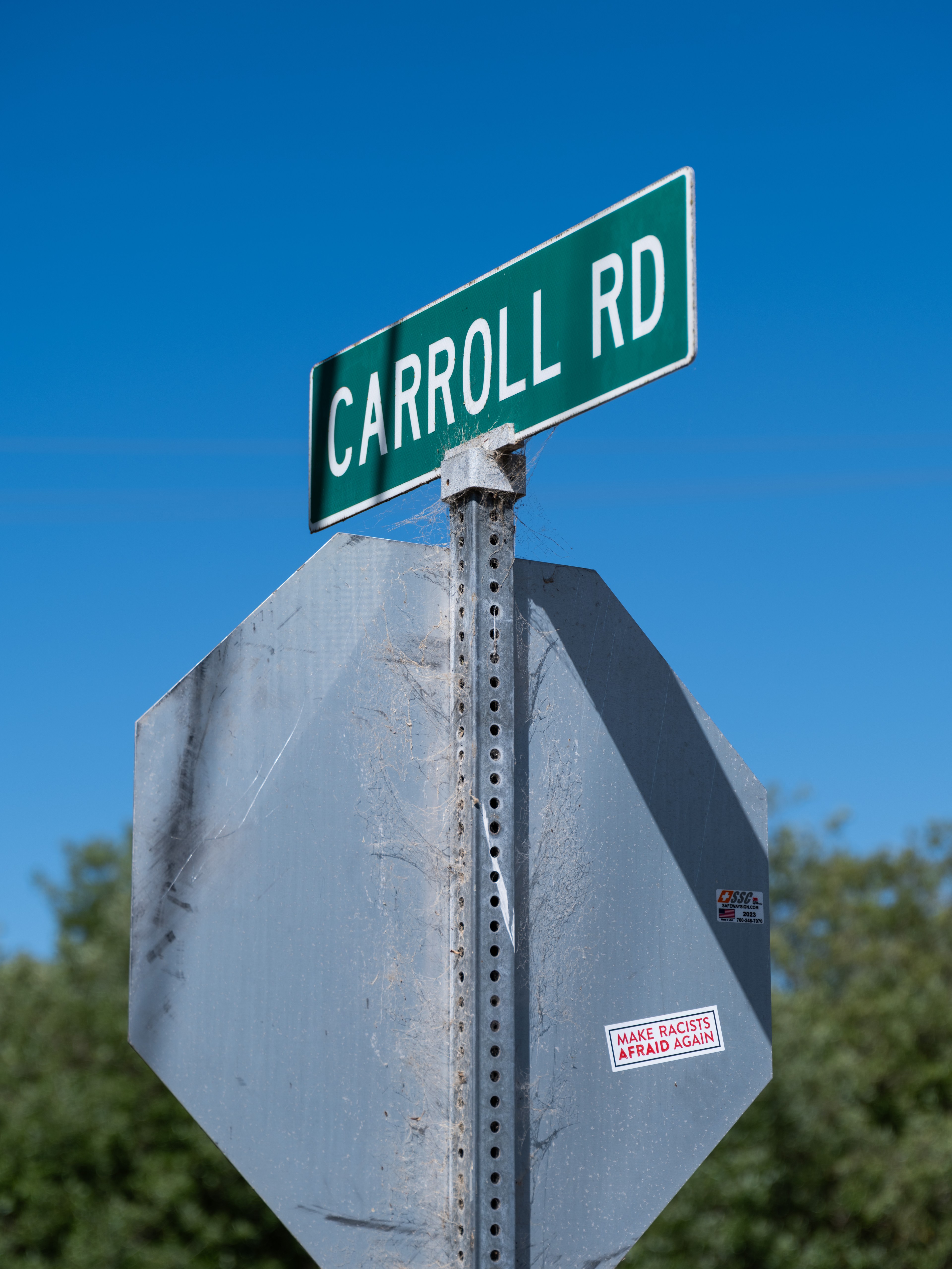 A street sign labeled &quot;CARROLL RD&quot; stands above a worn stop sign with a sticker that says &quot;MAKE RACISTS AFRAID AGAIN,&quot; set against a clear blue sky.