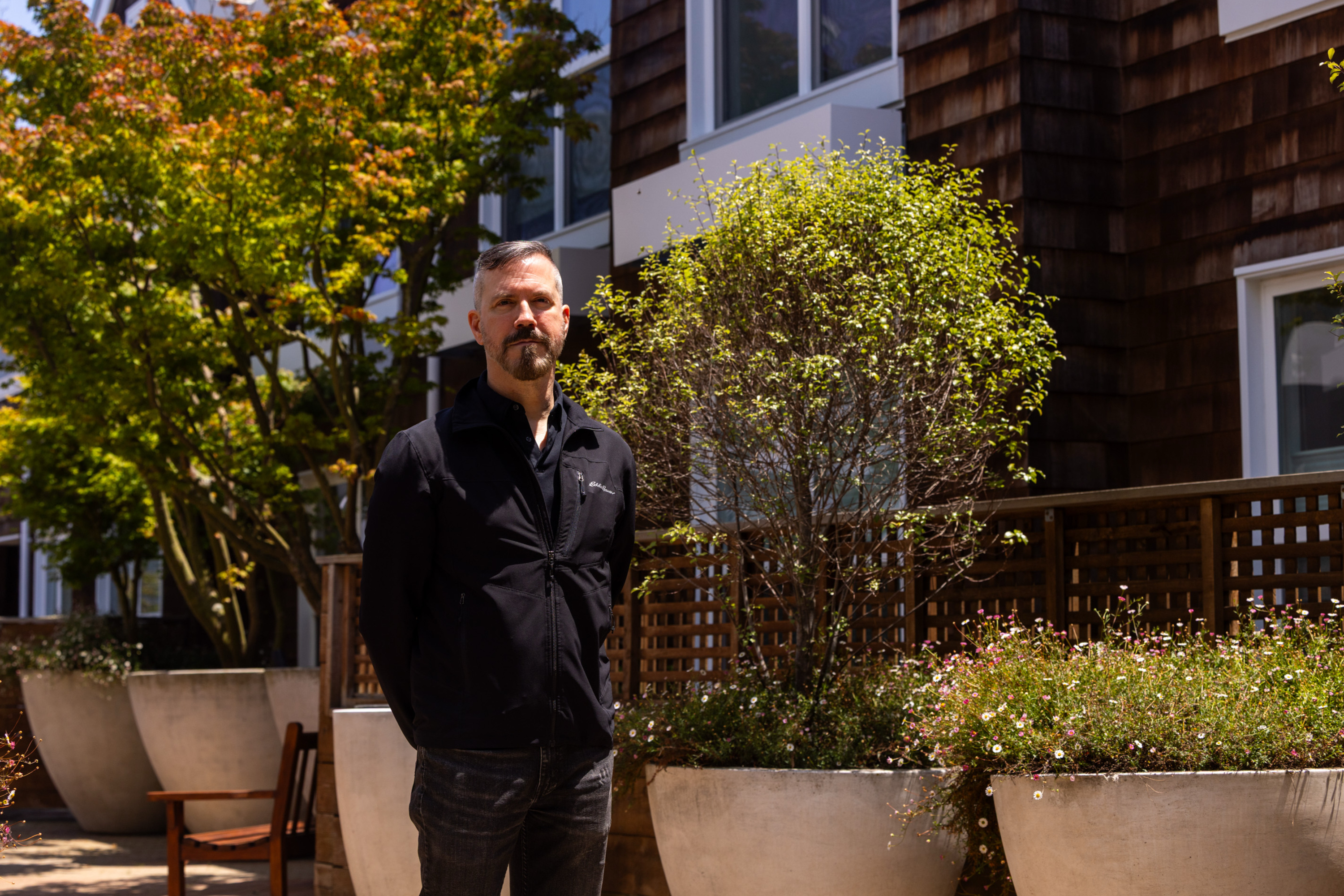 A person with short hair stands outdoors in front of a building, wearing a black jacket. Surrounding them are large planters with greenery and a tree.