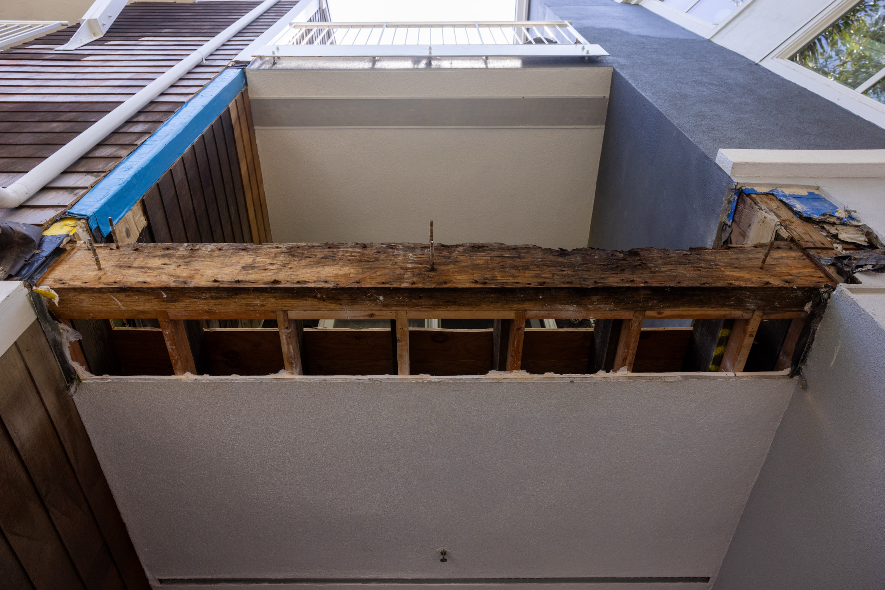 The image shows a partially demolished wooden beam structure under a small open area, framed by walls, with visible blue tape and exposed wood.