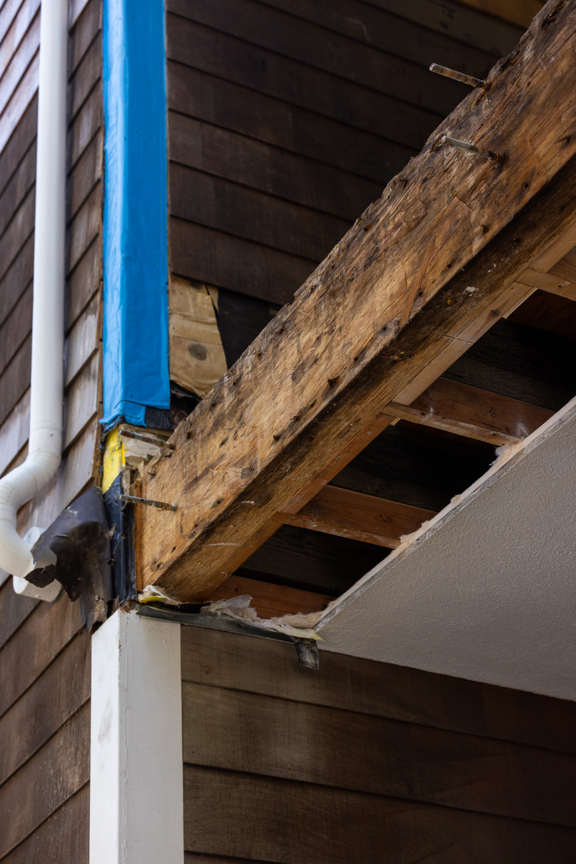 The image shows a corner of a building with damaged wood beams, a section covered by blue tape, and an exposed ceiling with missing plaster, revealing wooden beams.