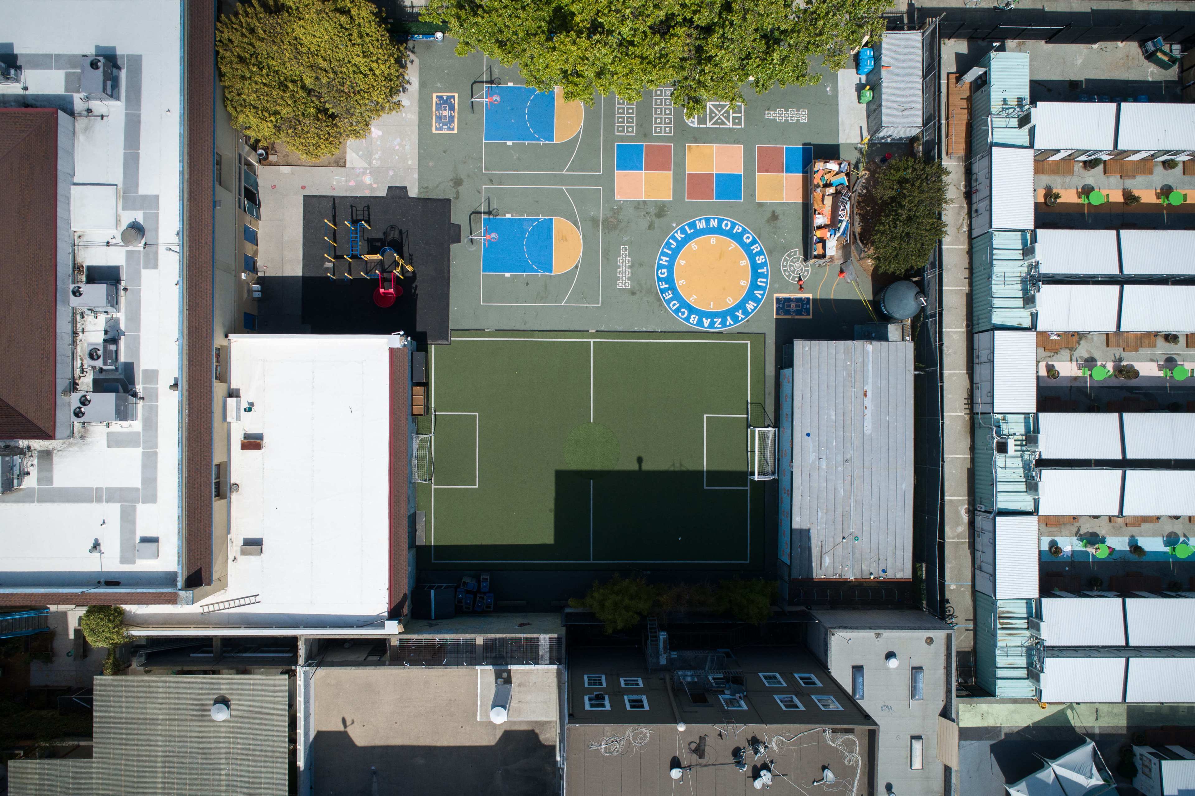 The image shows an aerial view of a colorful school playground with a basketball court, playground equipment, a small soccer field, and a seating area.