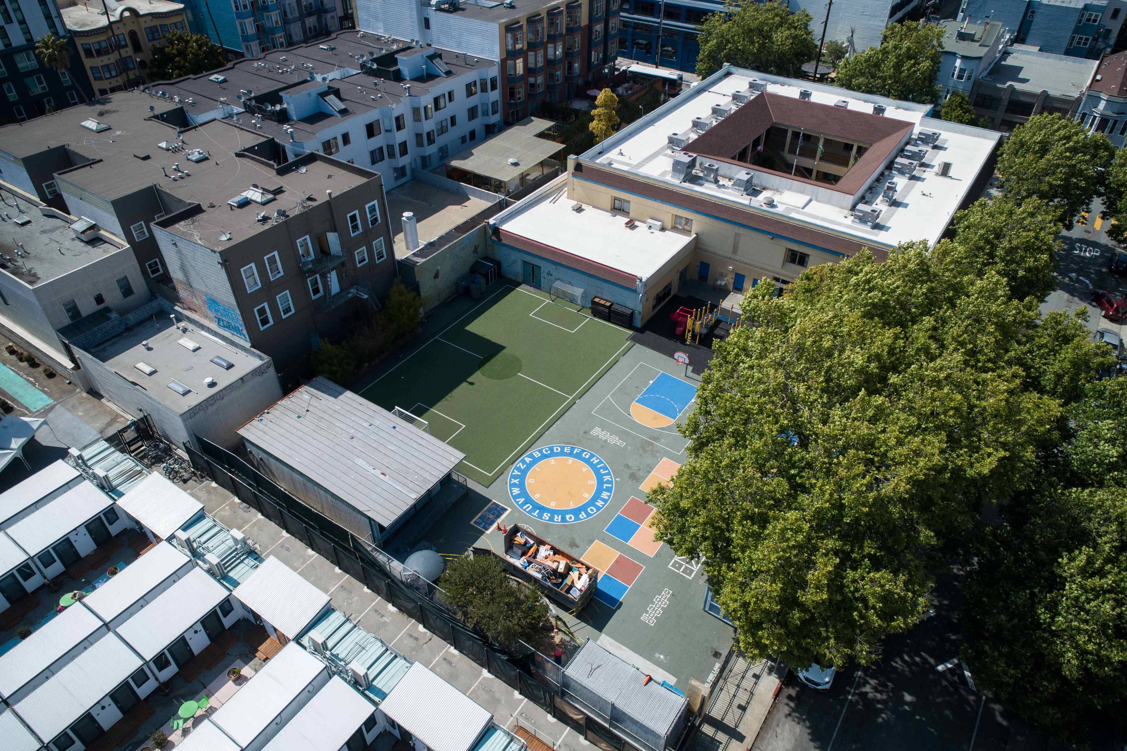 This aerial view shows a small urban schoolyard with a playground marked for different sports and activity areas. Surrounding buildings and trees are visible.