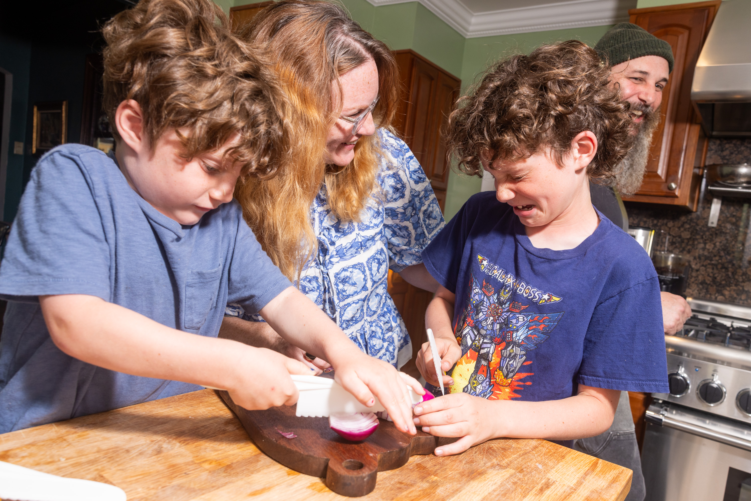 Two kids are cutting an onion on a wooden board in a kitchen, one looks like they are about to cry. Two adults are smiling and watching them.