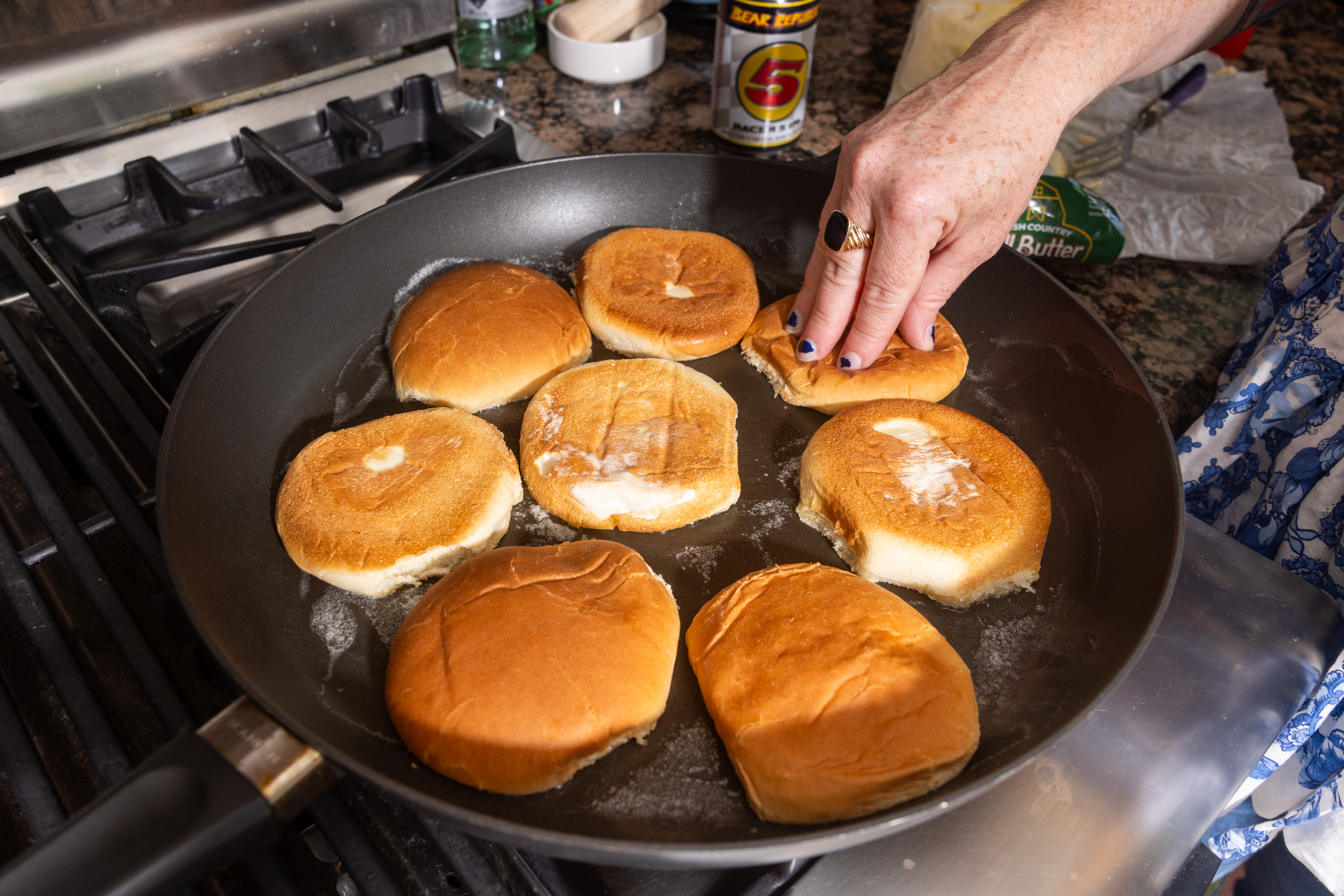 A hand is toasting eight hamburger bun halves in a frying pan on a stovetop, with butter melting on each one. A blue-patterned garment is partially visible.