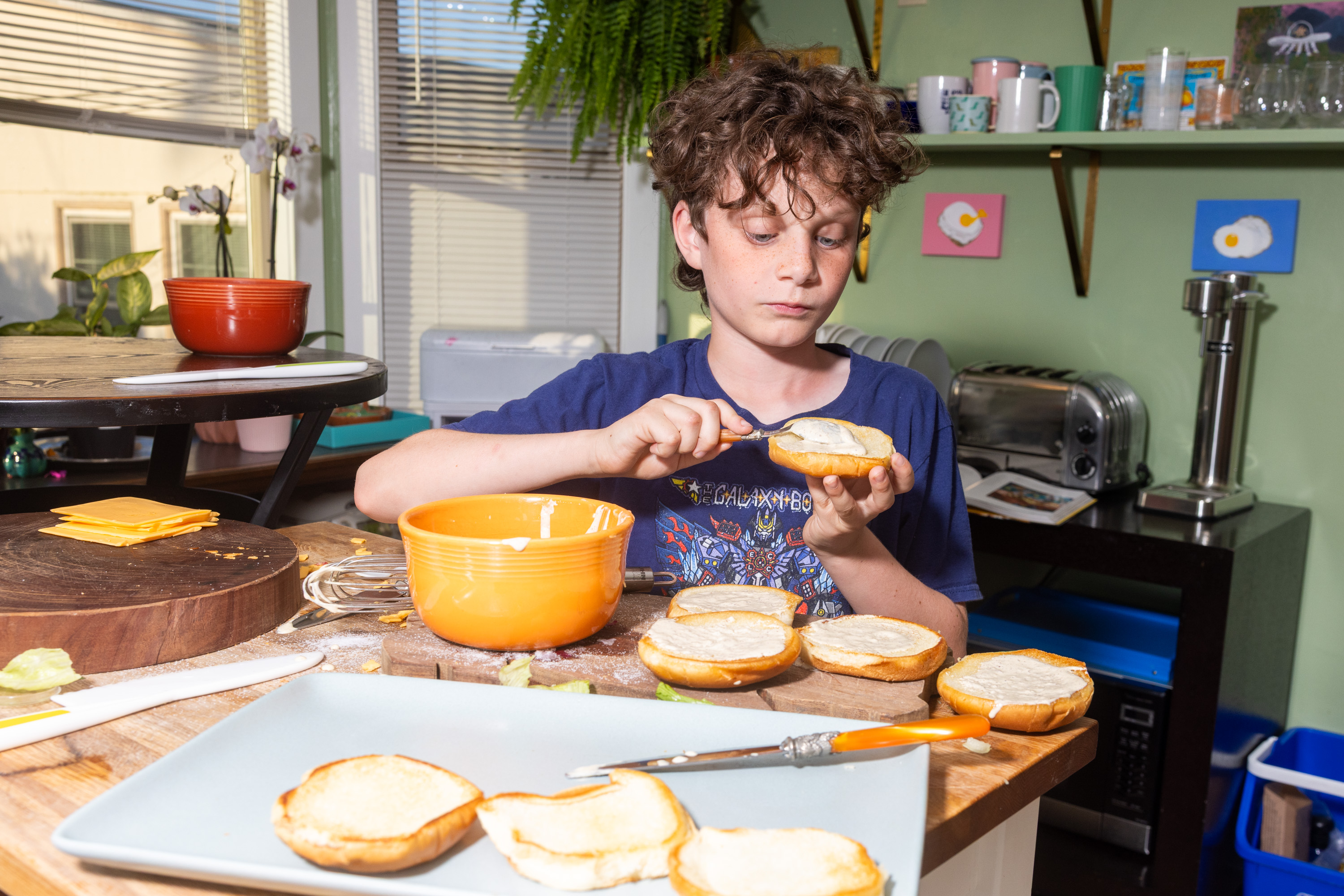 A child is spreading a creamy substance on buns in a kitchen. Surrounding them are a bowl, cheese slices, a toaster, and whimsical egg-themed art on the walls.