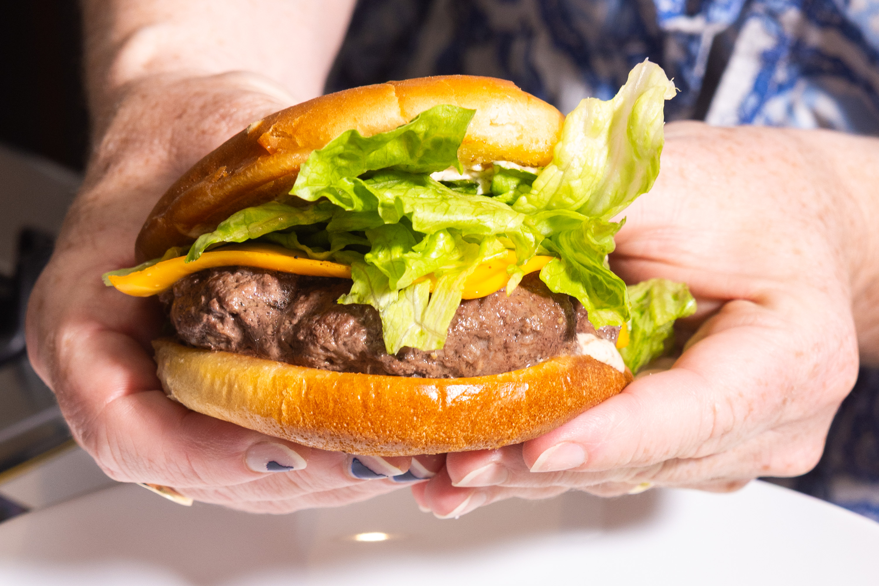 A person is holding a hamburger with a beef patty, cheddar cheese, and fresh lettuce between a shiny bun. The hands show nail art, and the background is blurred.