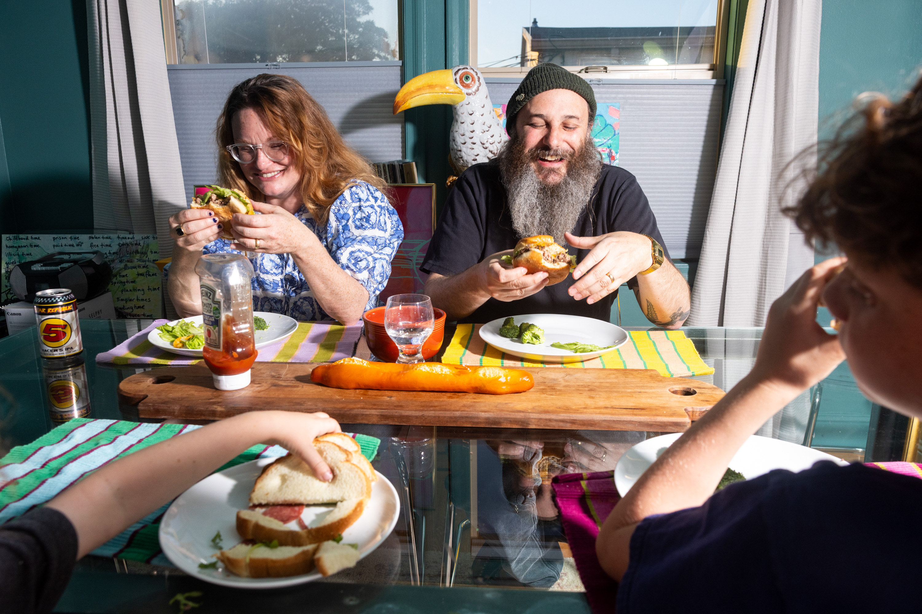 A family of three is joyfully eating sandwiches at a table. The table has a wooden platter, a bottle of ketchup, broccoli, and a can of soda.