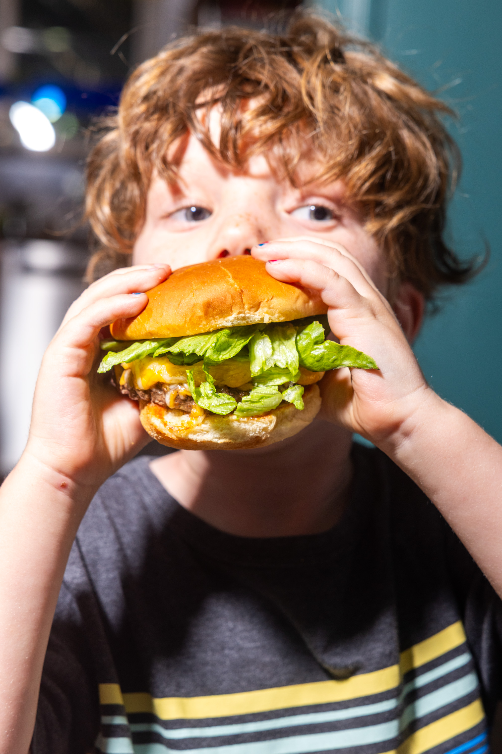 A child with curly hair takes a bite from a large burger with lettuce and cheese, using both hands to hold it. The child wears a striped shirt.