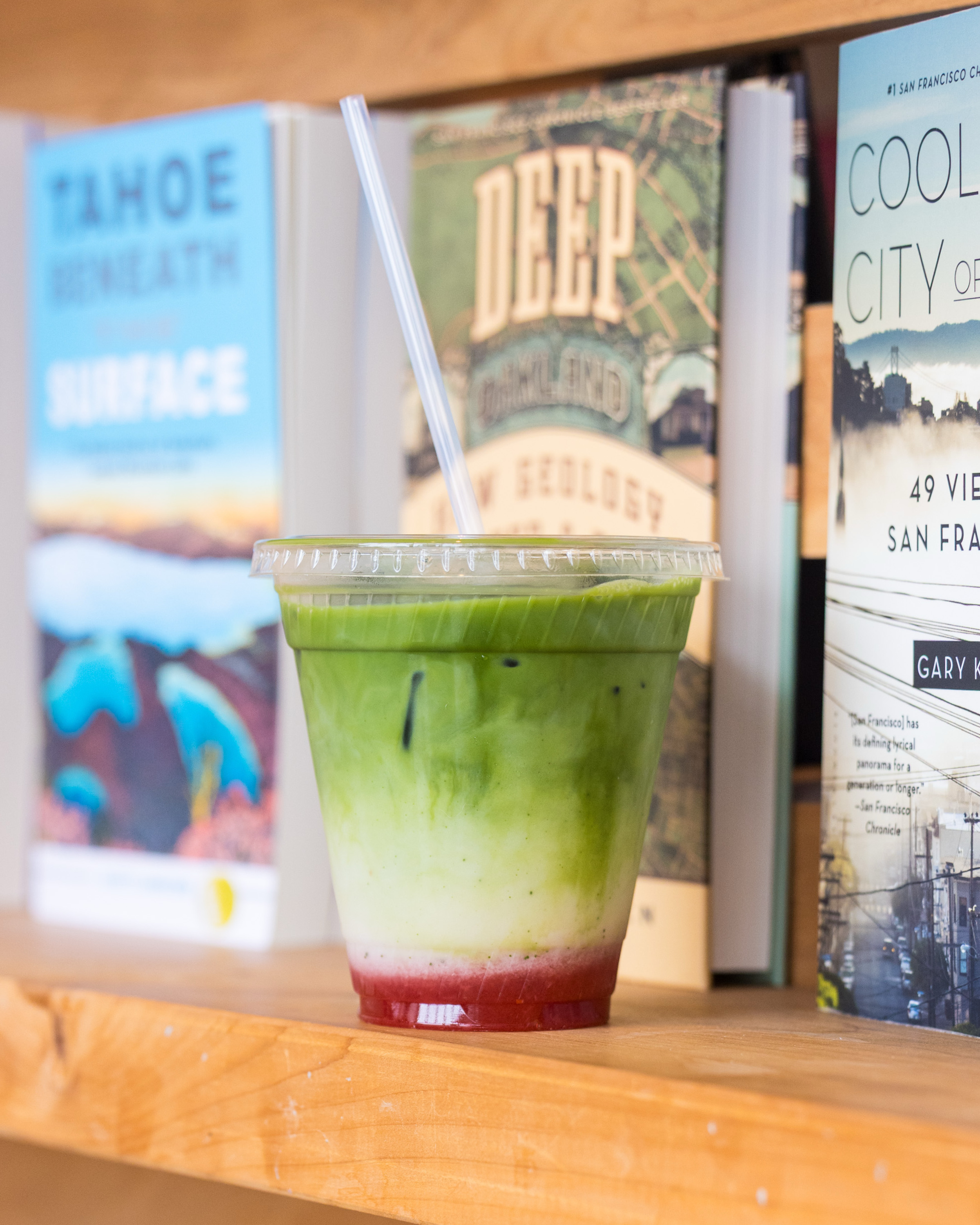 A green, white, and red layered smoothie in a plastic cup with a straw sits on a wooden shelf between three books with colorful covers.